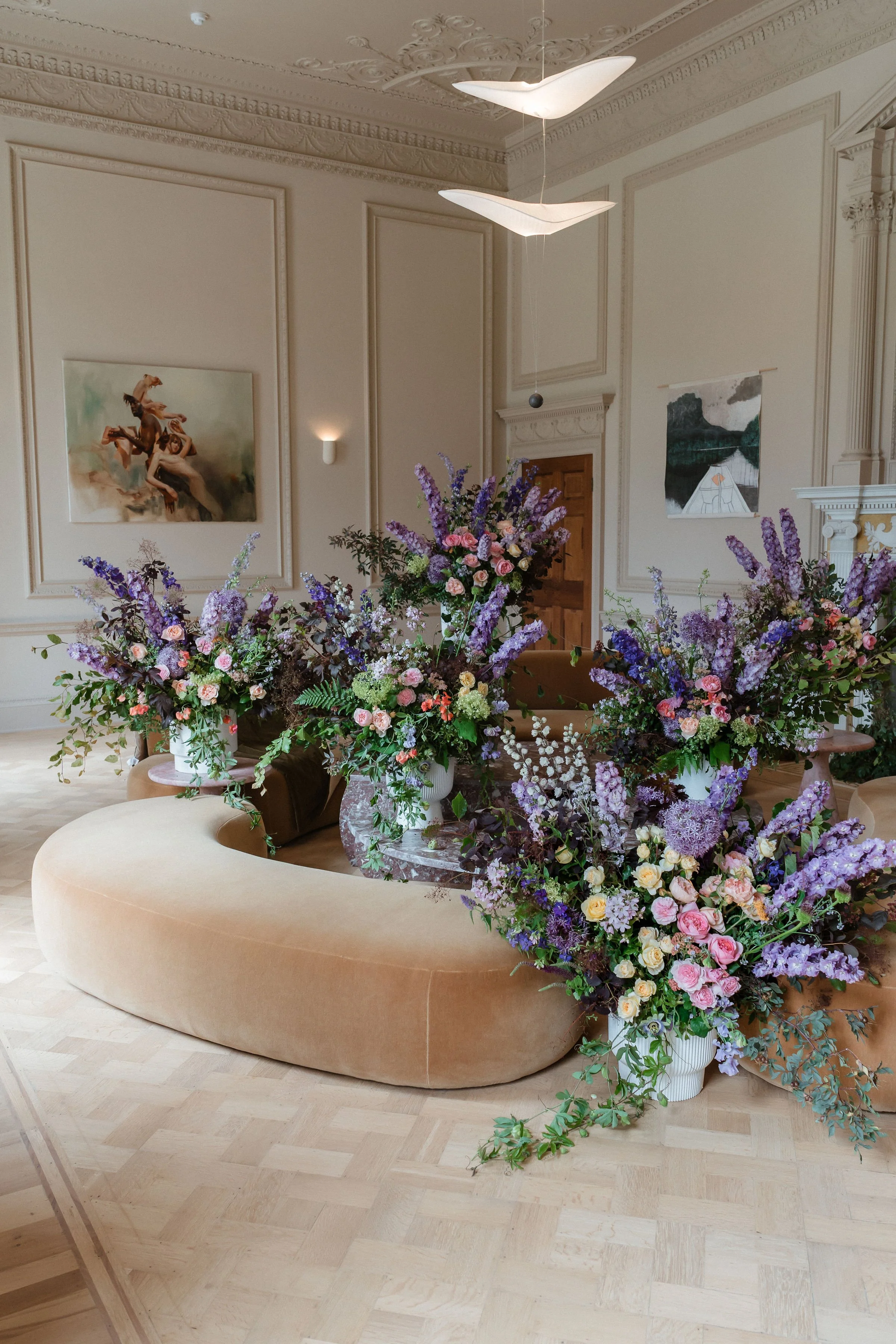 Elegant room at Denton Reserve decorated with multiple large flower arrangements featuring purple, pink, and white flowers, placed on a cream-coloured curved sofa in front of a wooden door and framed artwork on the walls.