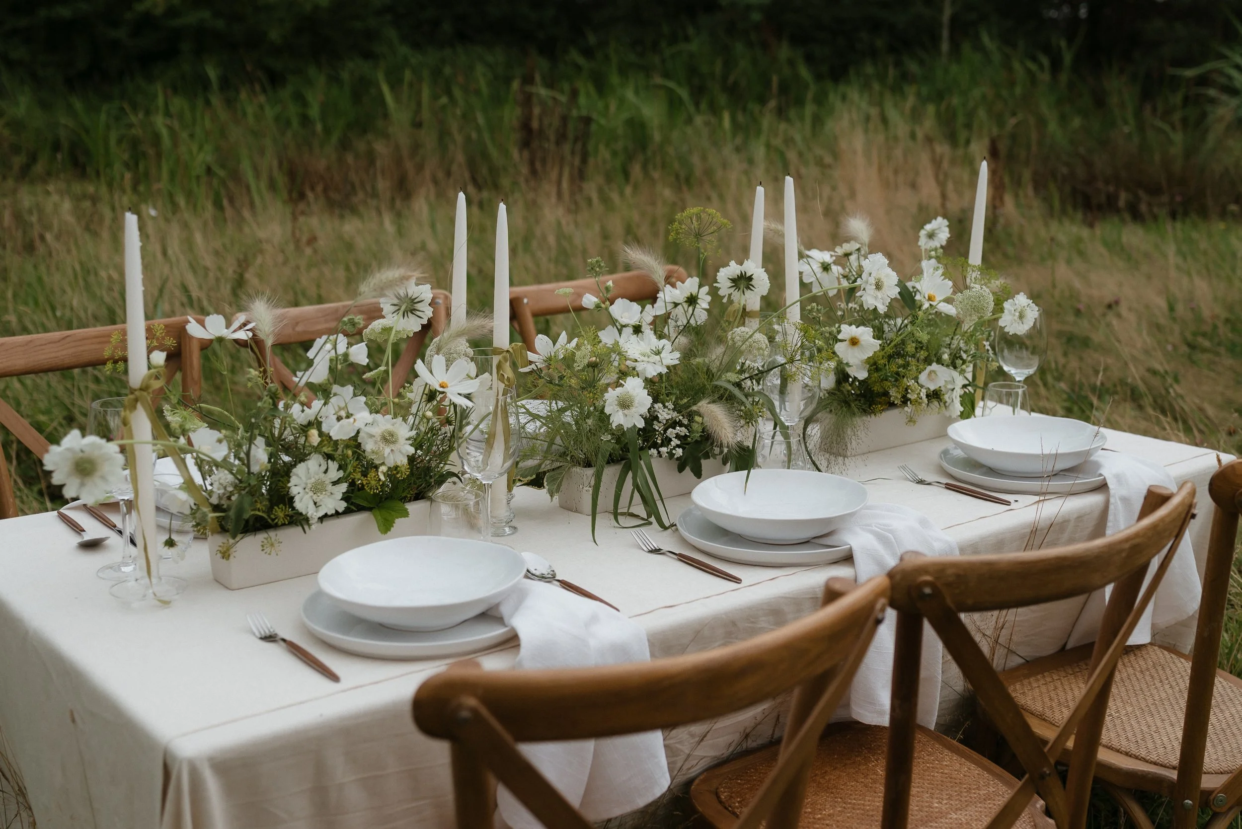 Outdoor dining table decorated with white flowers, candles, and tableware, set in a natural grassy area.