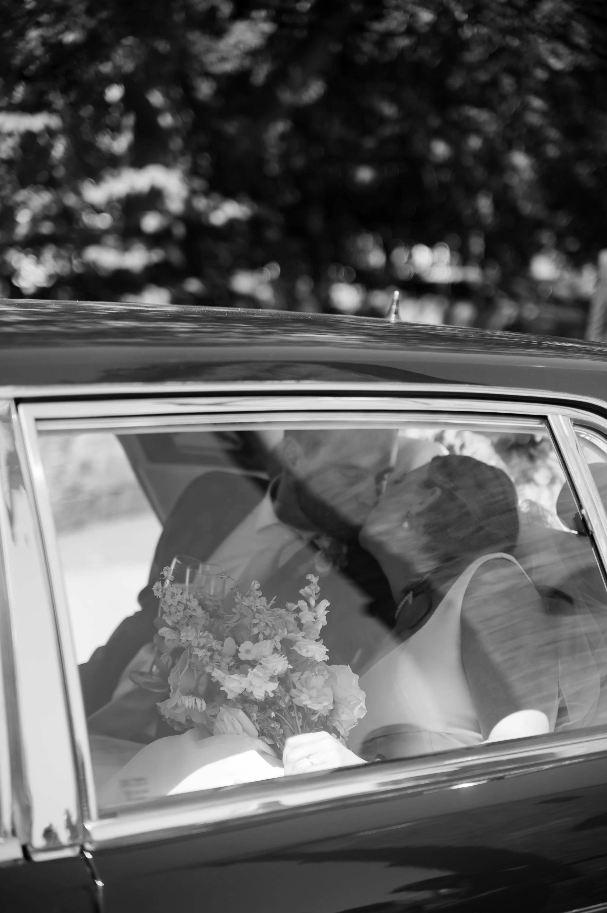 A black-and-white photo of a couple sitting inside a car, sharing a kiss. The woman is holding a bouquet of flowers, and the car has a classic style. Trees are visible outside the car window.