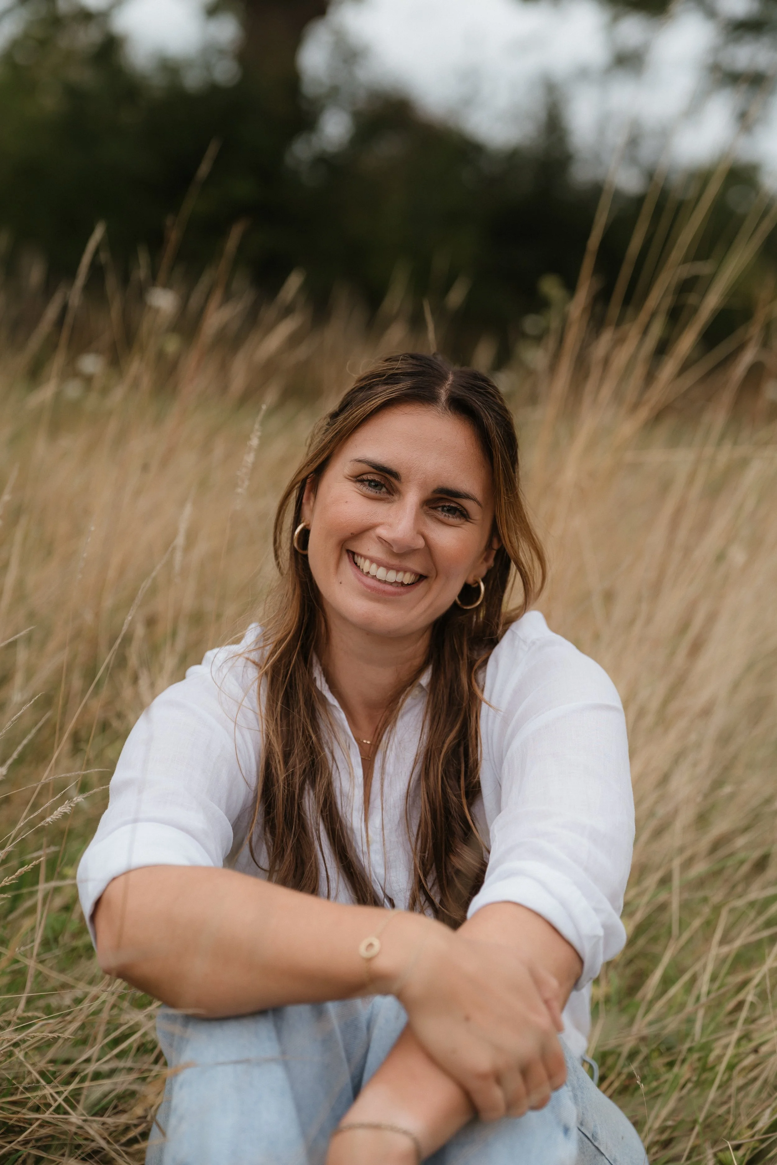 A woman with long brown hair, wearing a white shirt and blue jeans, sitting in a field of tall grass, smiling at the camera.