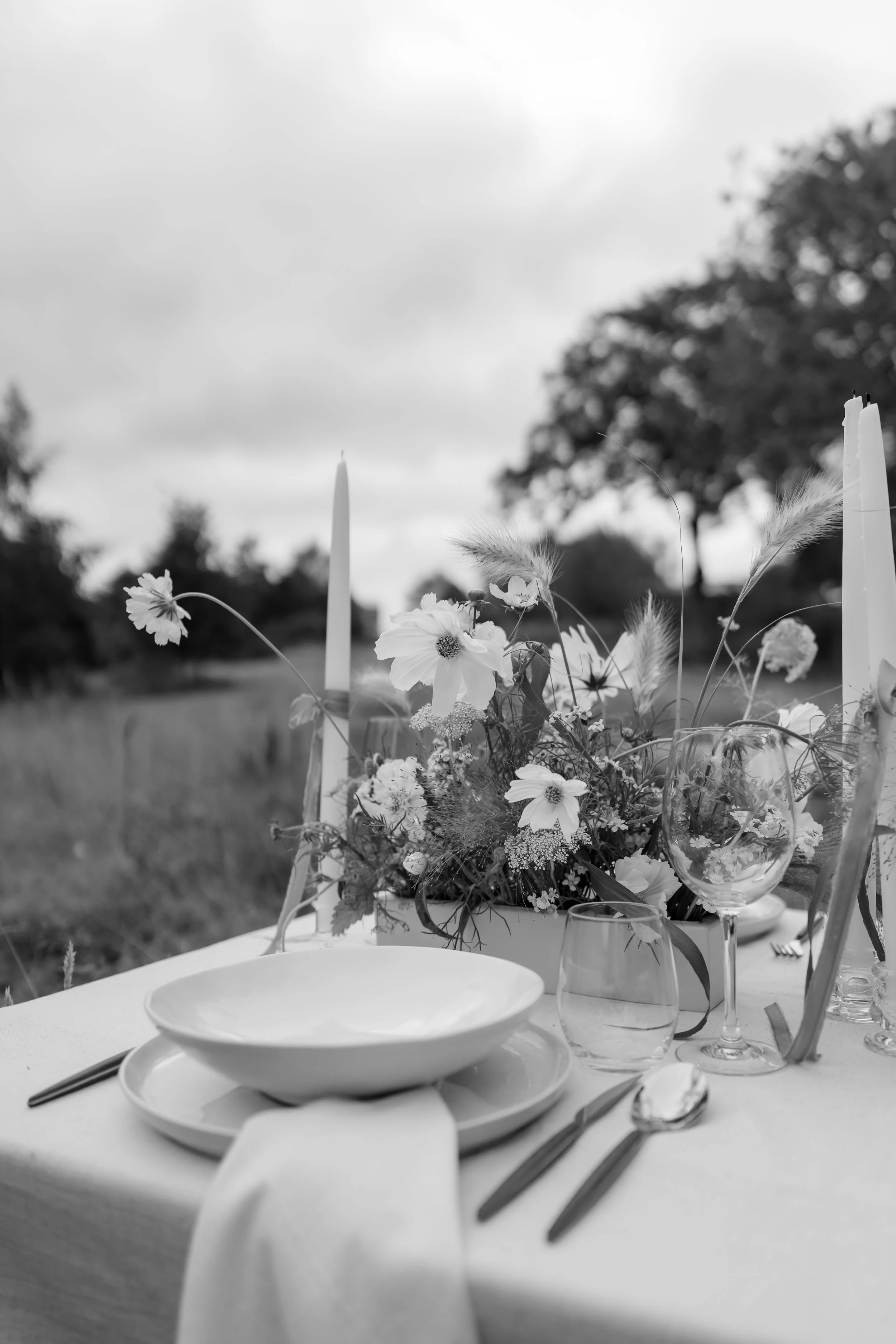A black and white photo of an outdoor dining table set with plates, glasses, utensils, a floral centerpiece, and tall candles, against a backdrop of trees and cloudy sky.