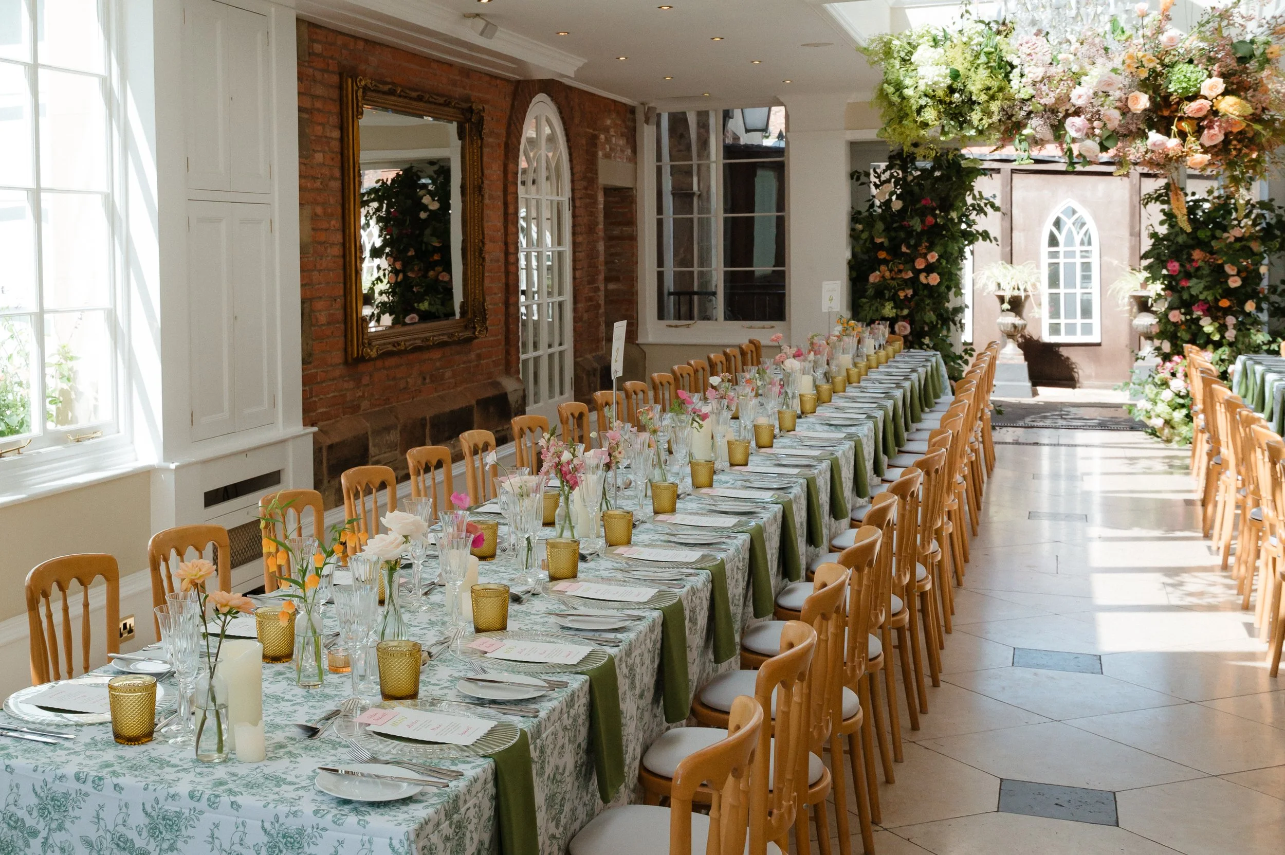 Long dining table set for an event with floral arrangements, candles, and place settings, inside a bright room with large windows and brick walls at Goldsborough Hall.