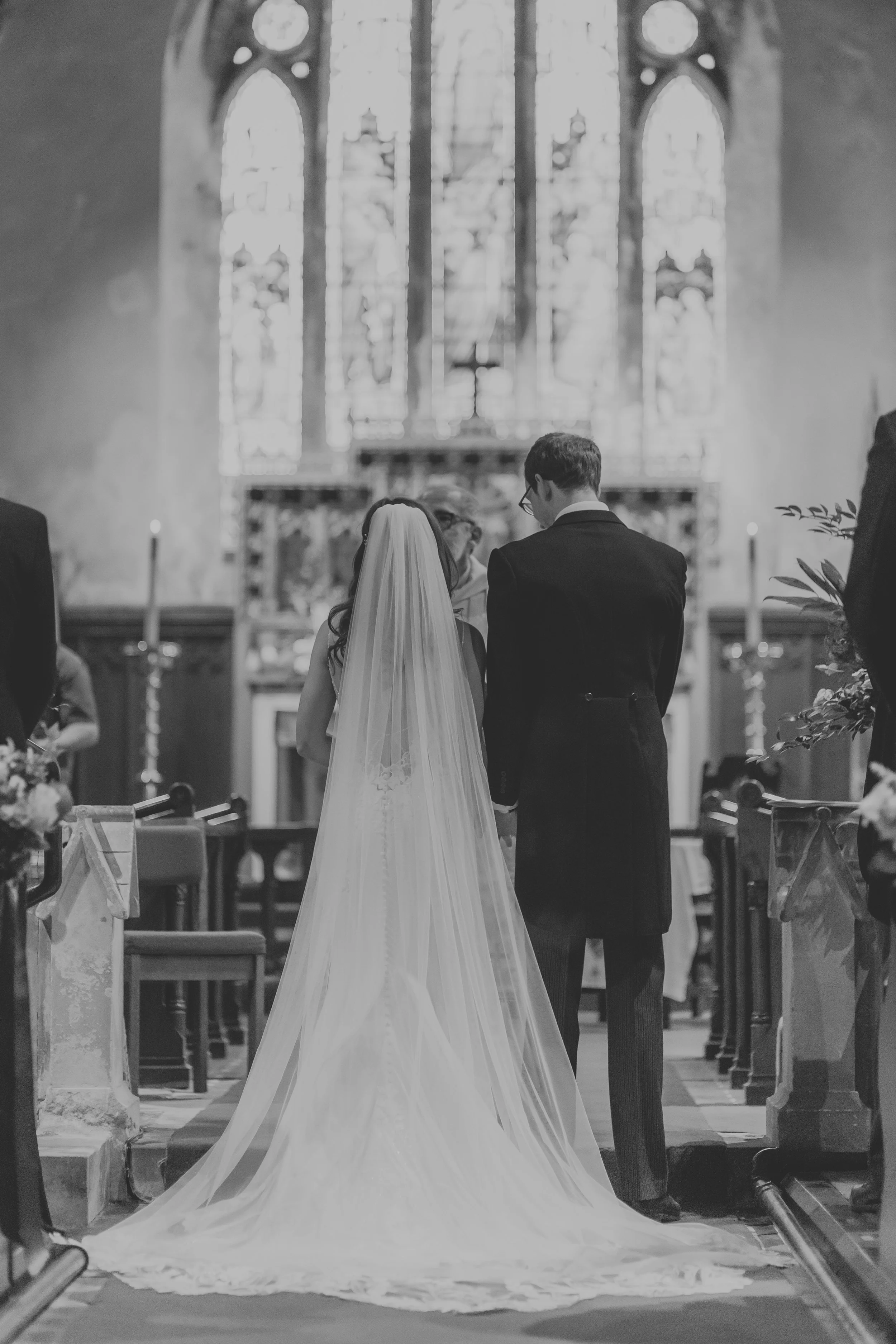 A black and white photo of a bride and groom standing at the altar in a church next to Goldsborough Hall during their wedding ceremony, with a priest officiating.