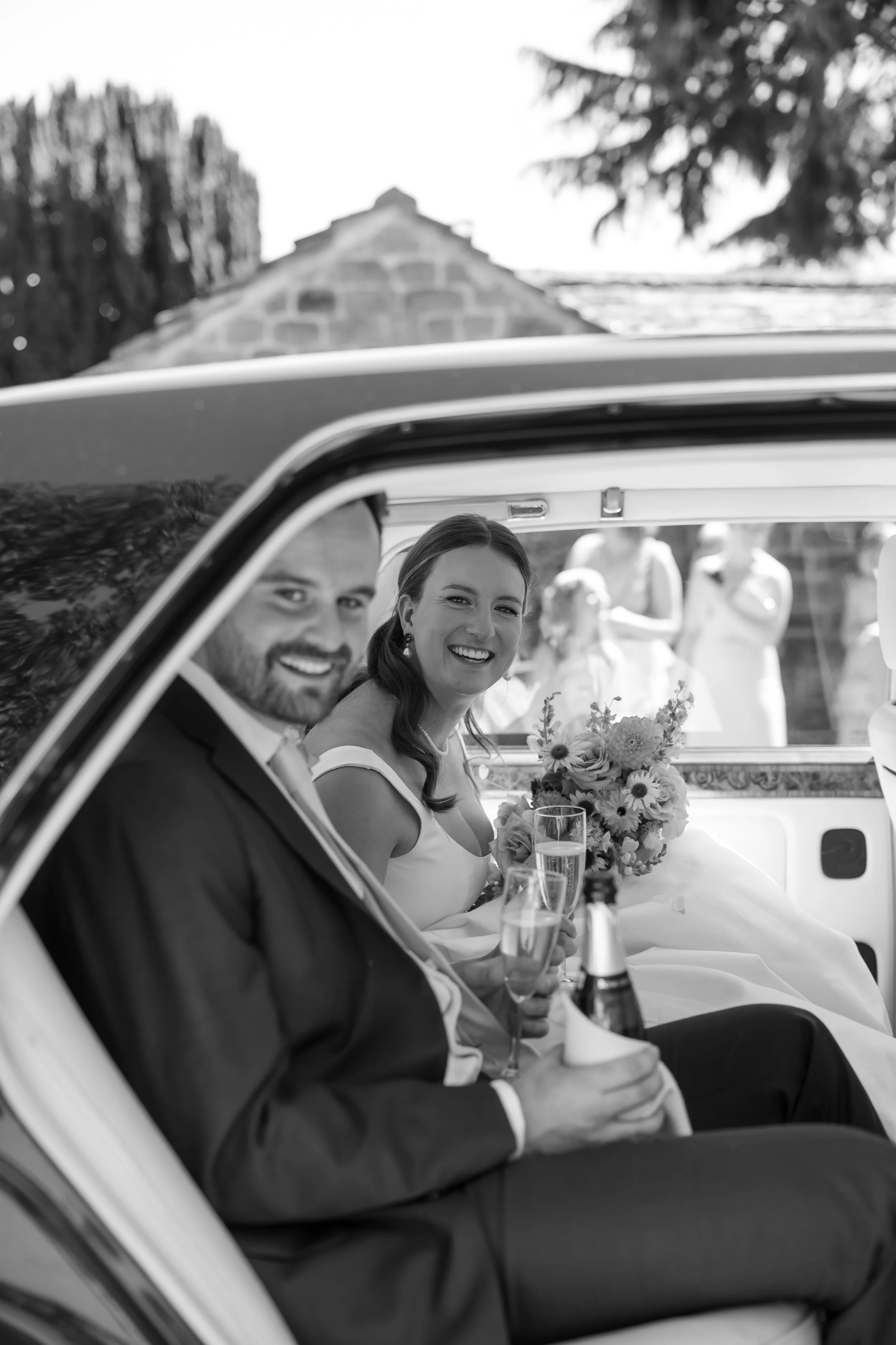 Black and white photo of a smiling bride and groom sitting inside a vintage car during their wedding celebration, holding champagne glasses and a bouquet of flowers.