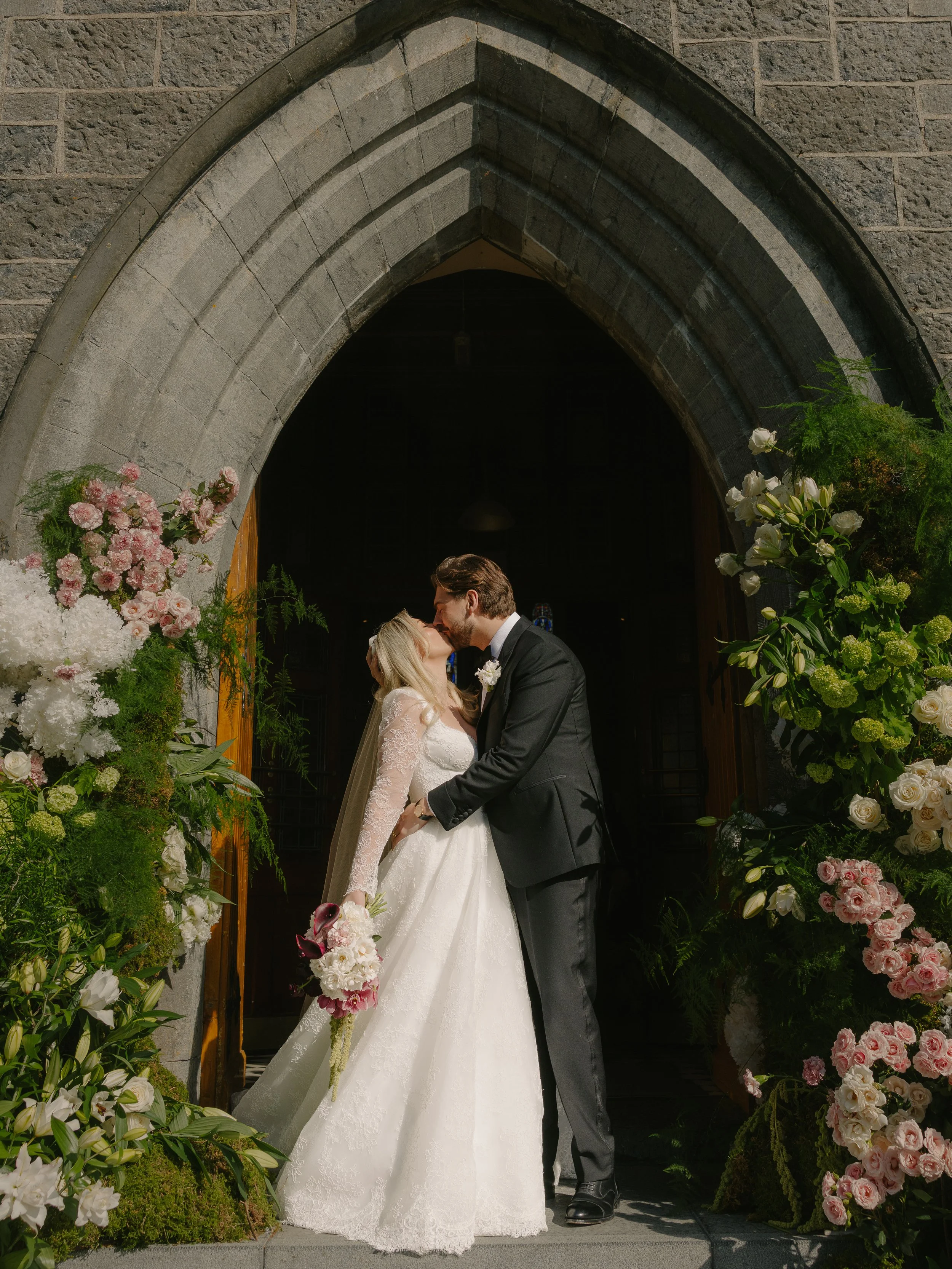 A bride and groom stand in the church door way with big green, white and pink floral columns framing them as they share a kiss after their wedding ceremony.