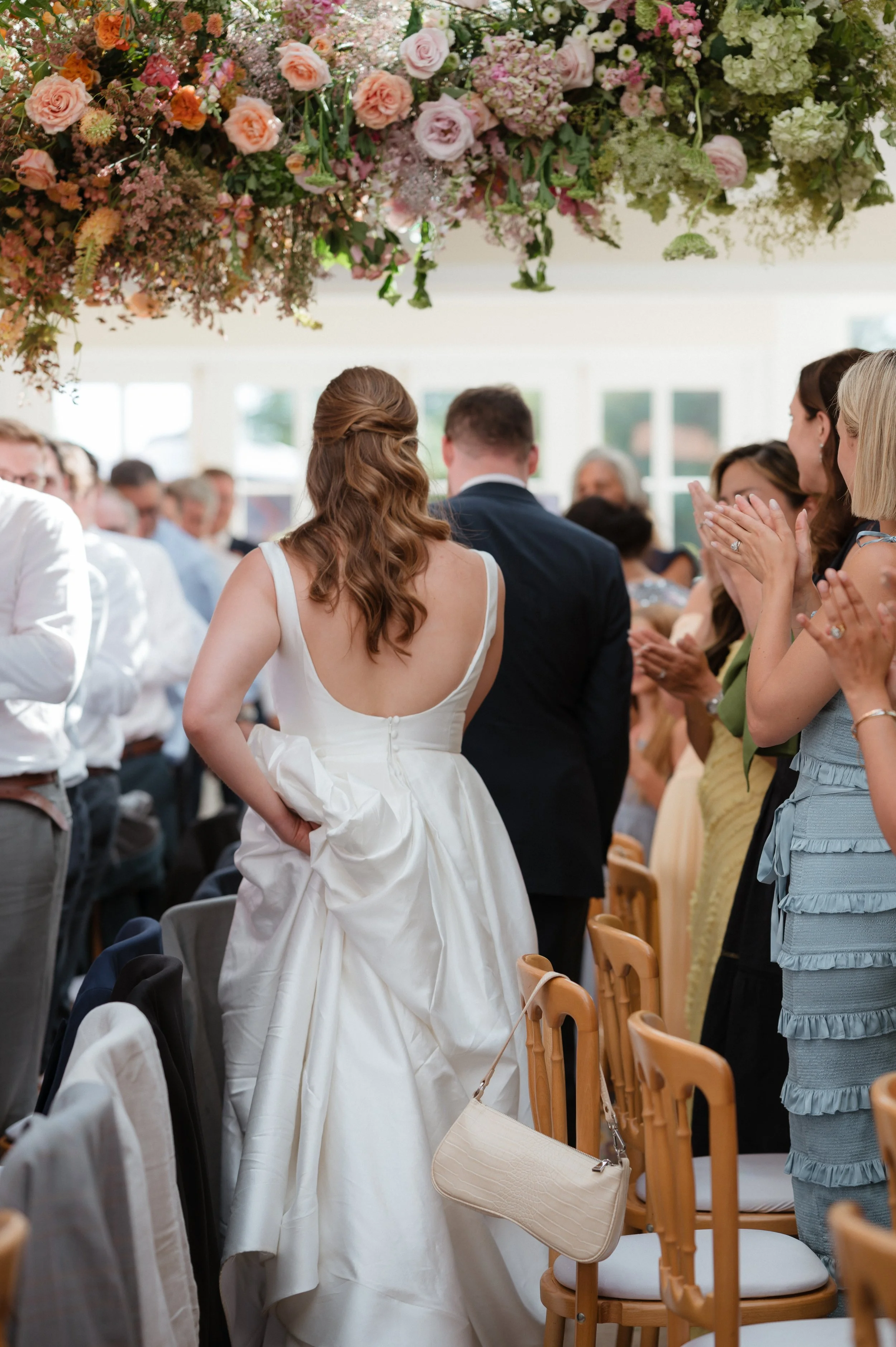 A bride in a white wedding dress with a low back, holding a jacket, standing among seated wedding guests in a decorated reception hall at Goldsborough Hallwith a large floral arrangement overhead.