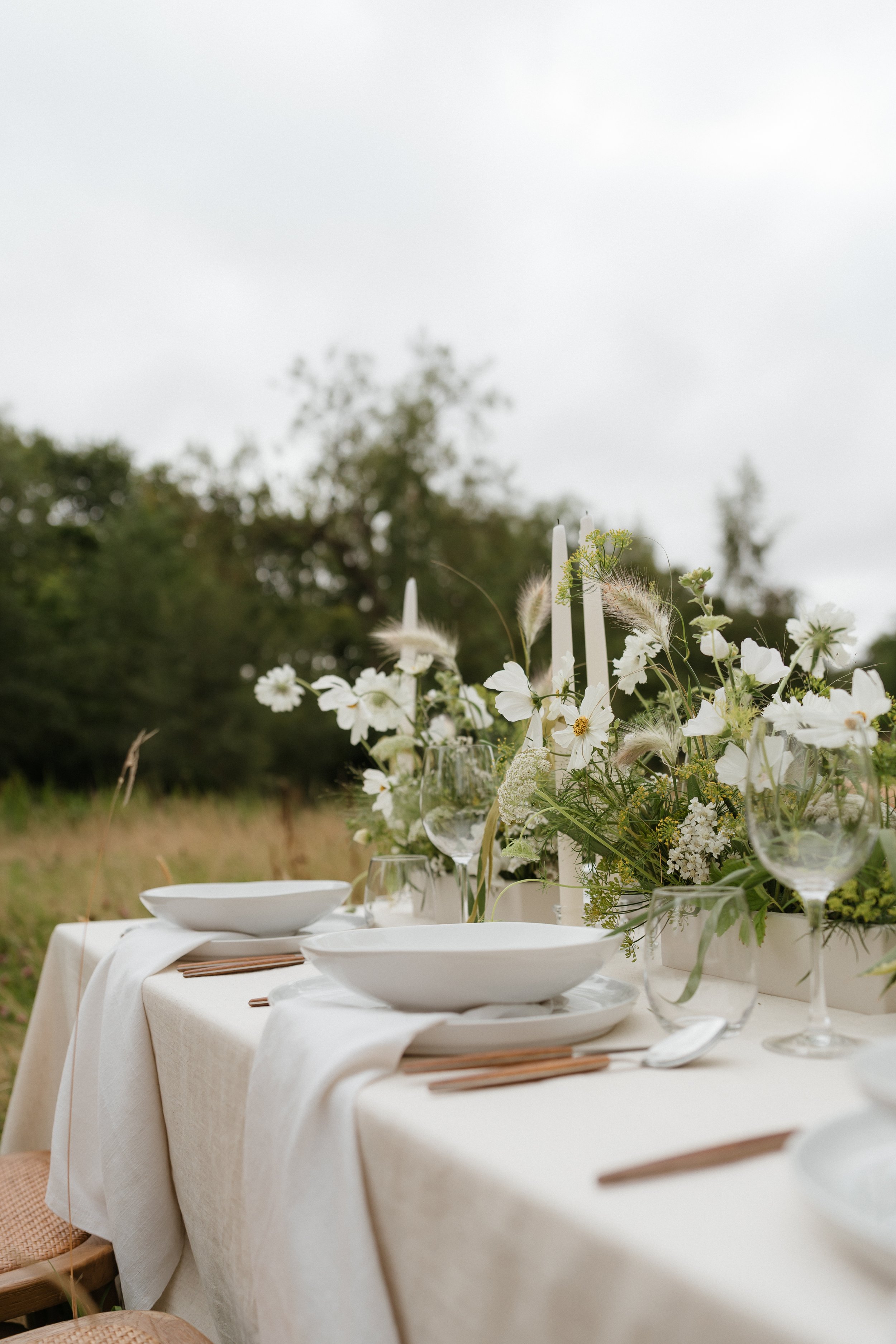Elegant outdoor dining table set with white tablecloth, white plates, glassware, wooden utensils, and a floral centerpiece with white flowers and greenery, in a natural outdoor setting under an overcast sky.