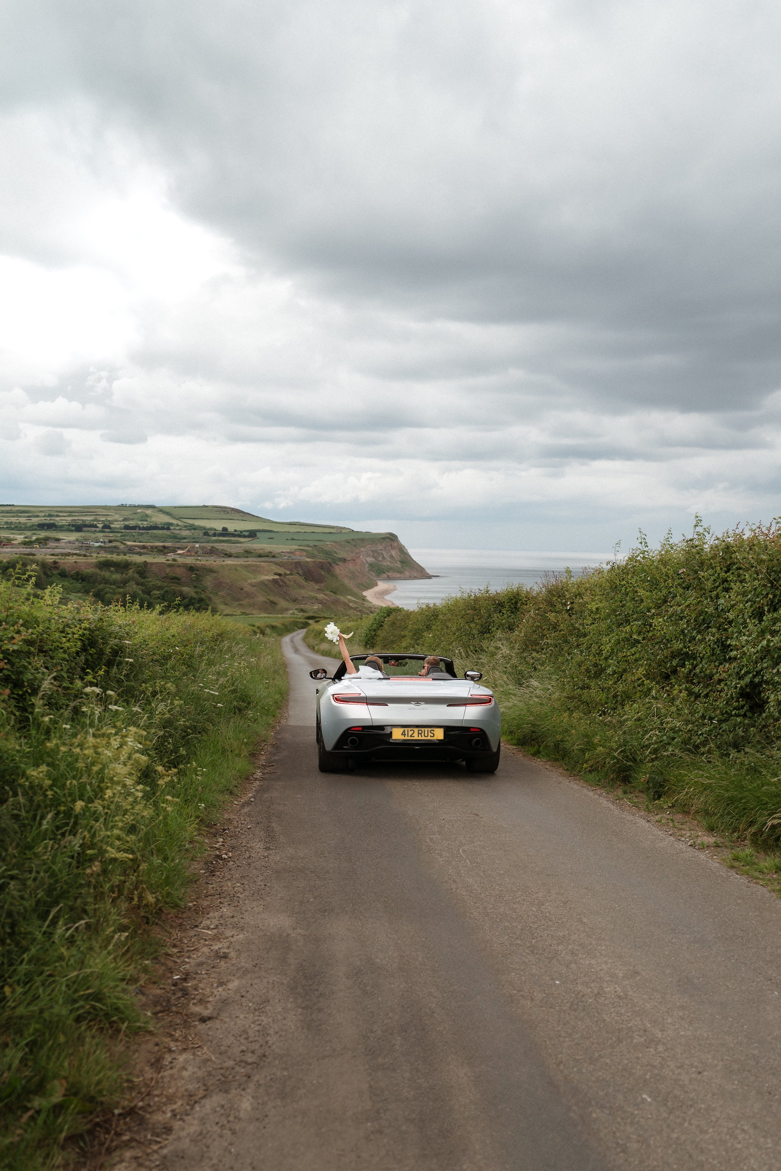 A wedding couple in a silver convertible car on a rural road with Yorkshire coastline in the background. The bride is holding a bouquet and raising her arm.