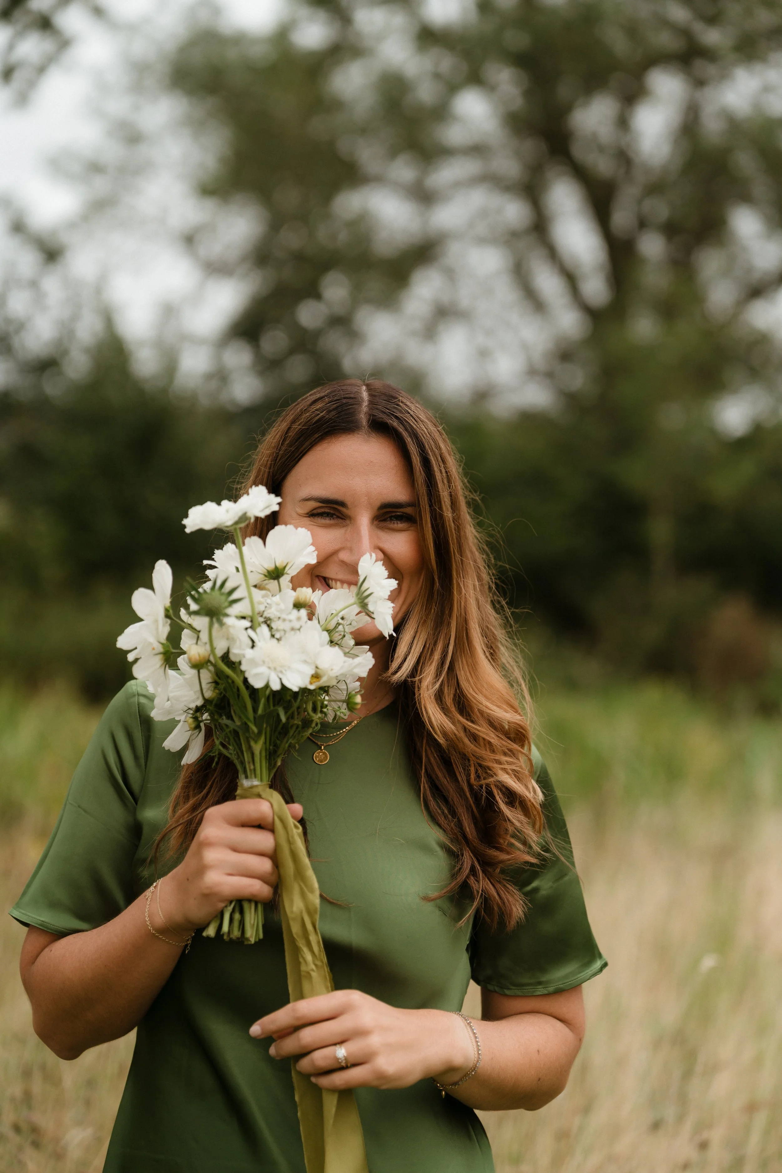 A woman with long brown hair, wearing a green dress, holding a bouquet of white flowers, smiling outdoors in a natural setting.