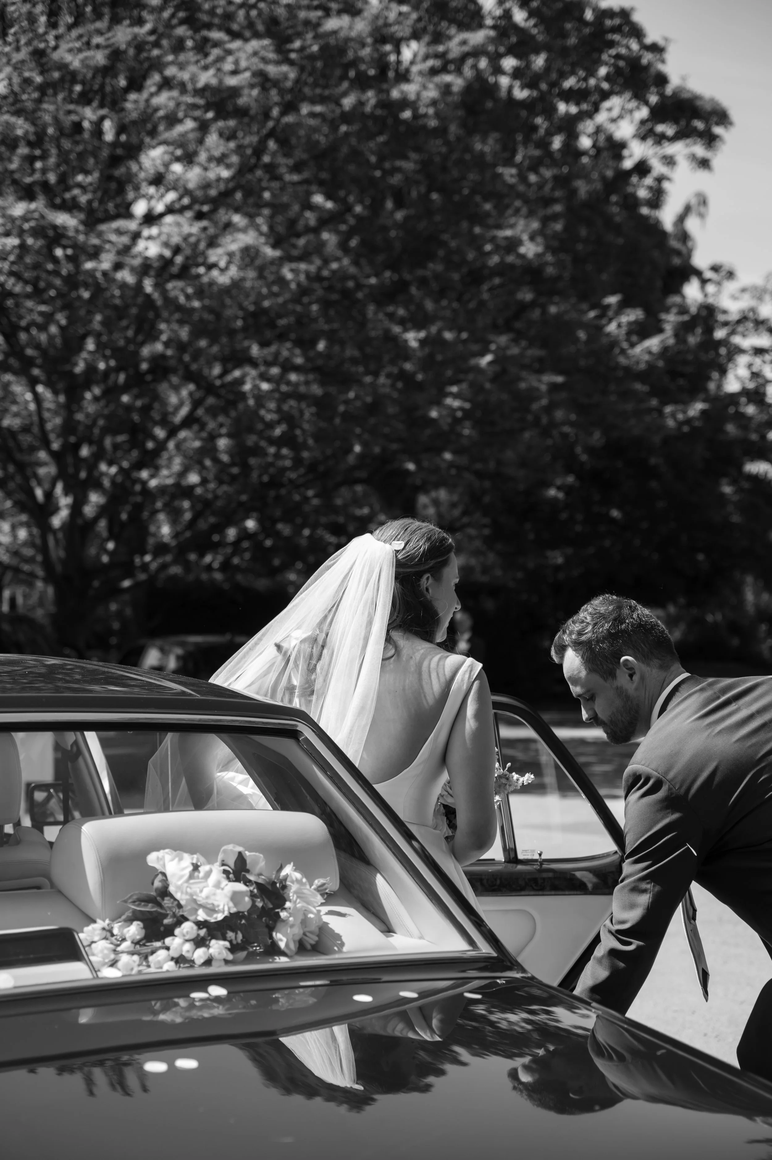 A bride in a wedding dress and veil is sitting in a car, with a man leaning in, outdoors on a sunny day.