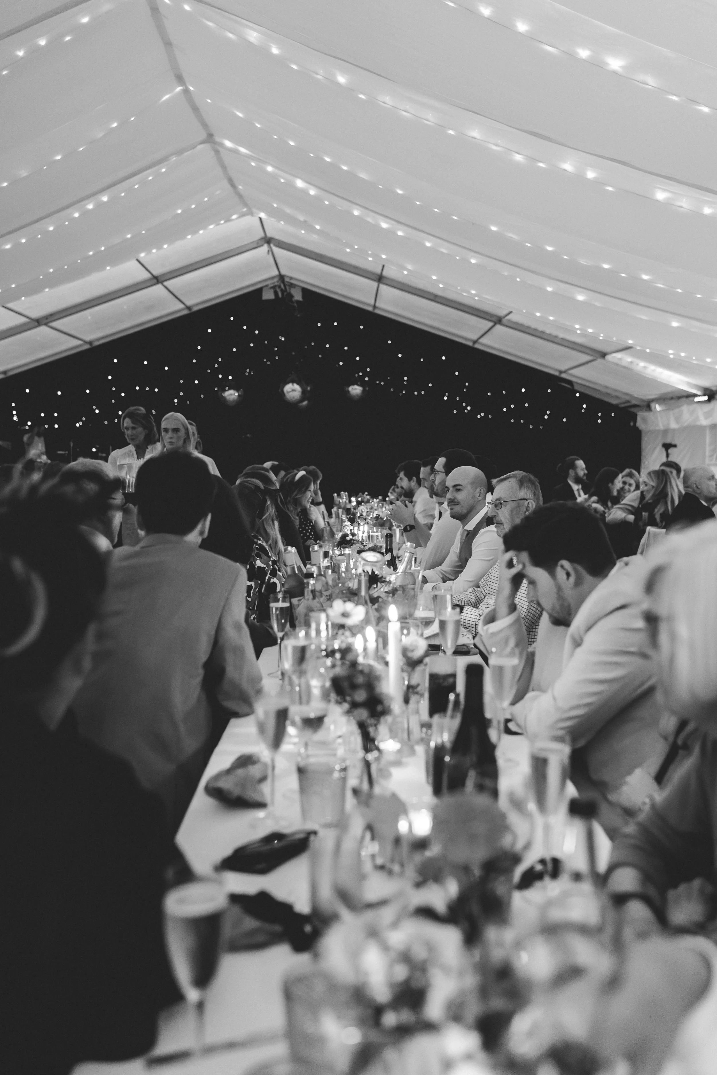 Black and white photo of a long banquet table inside a decorated marquee in Cheshire with string lights, amid guests seated and socialising.