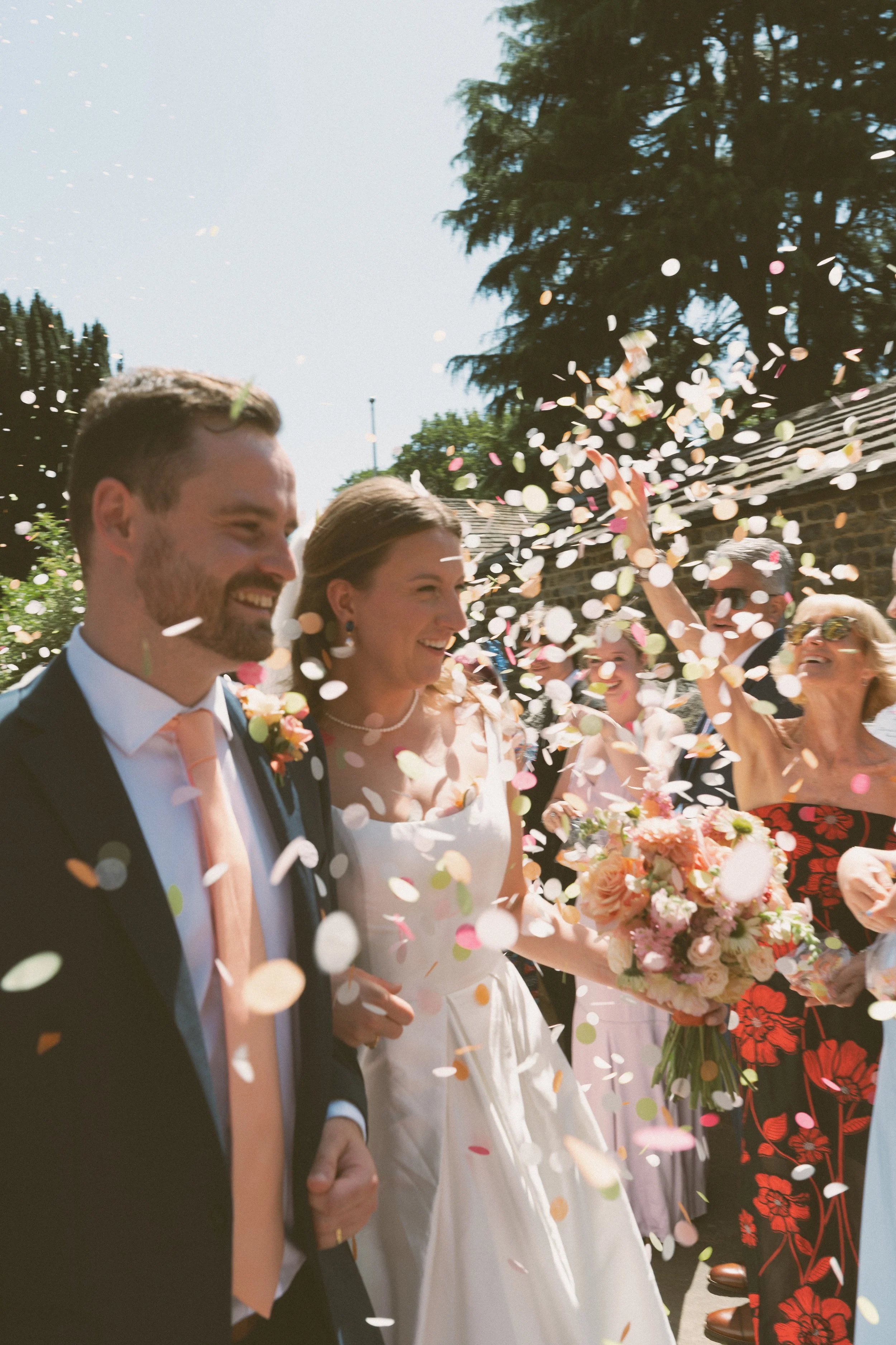 A wedding celebration outdoors on a sunny day with a bride in a white dress, groom in a dark suit, and guests throwing colorful confetti. The bride holds a bouquet, and people are smiling and enjoying the moment.