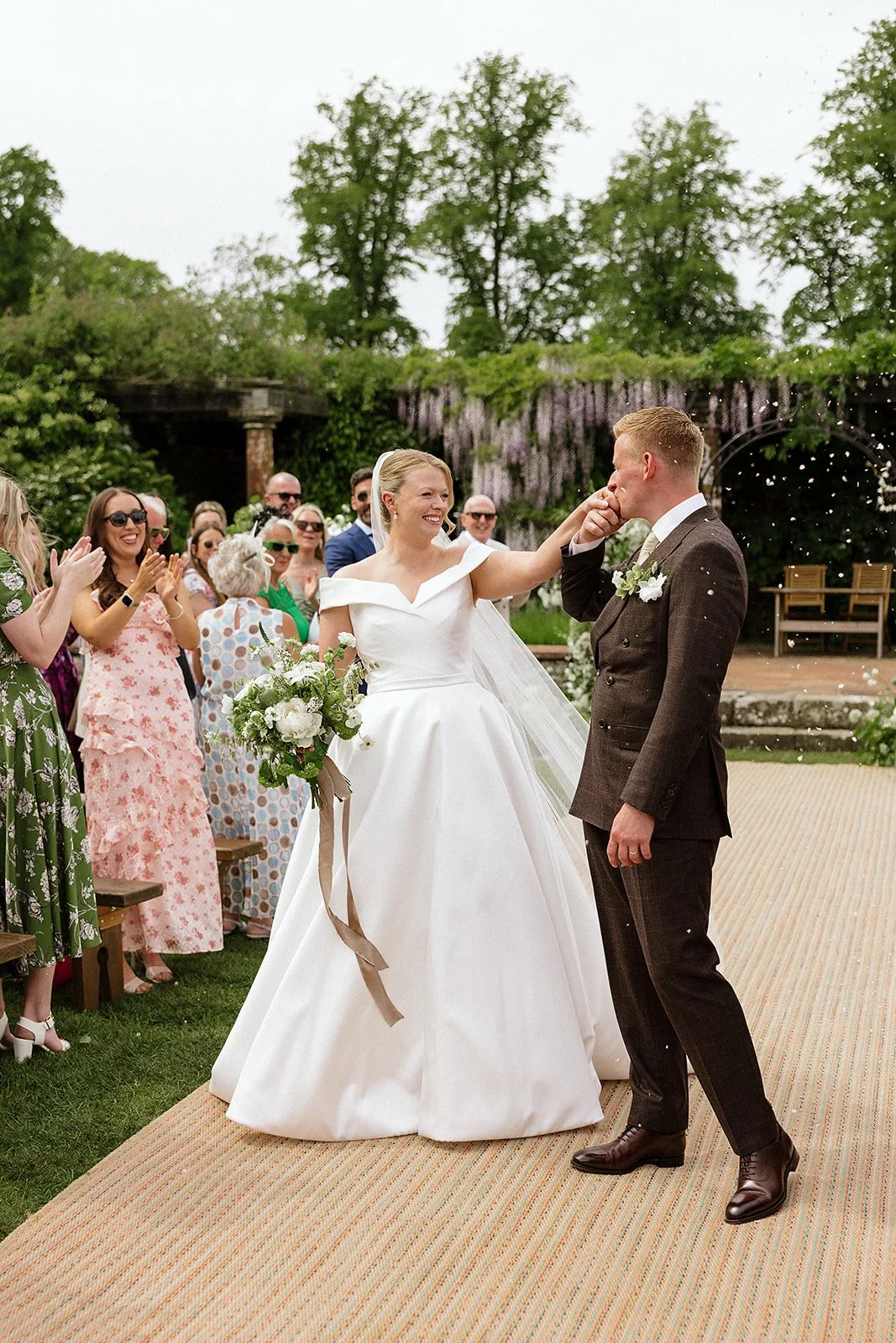 A bride and groom celebrate as they walk down the aisle. Confetti canons go off around them and the groom kisses the bride's hand.