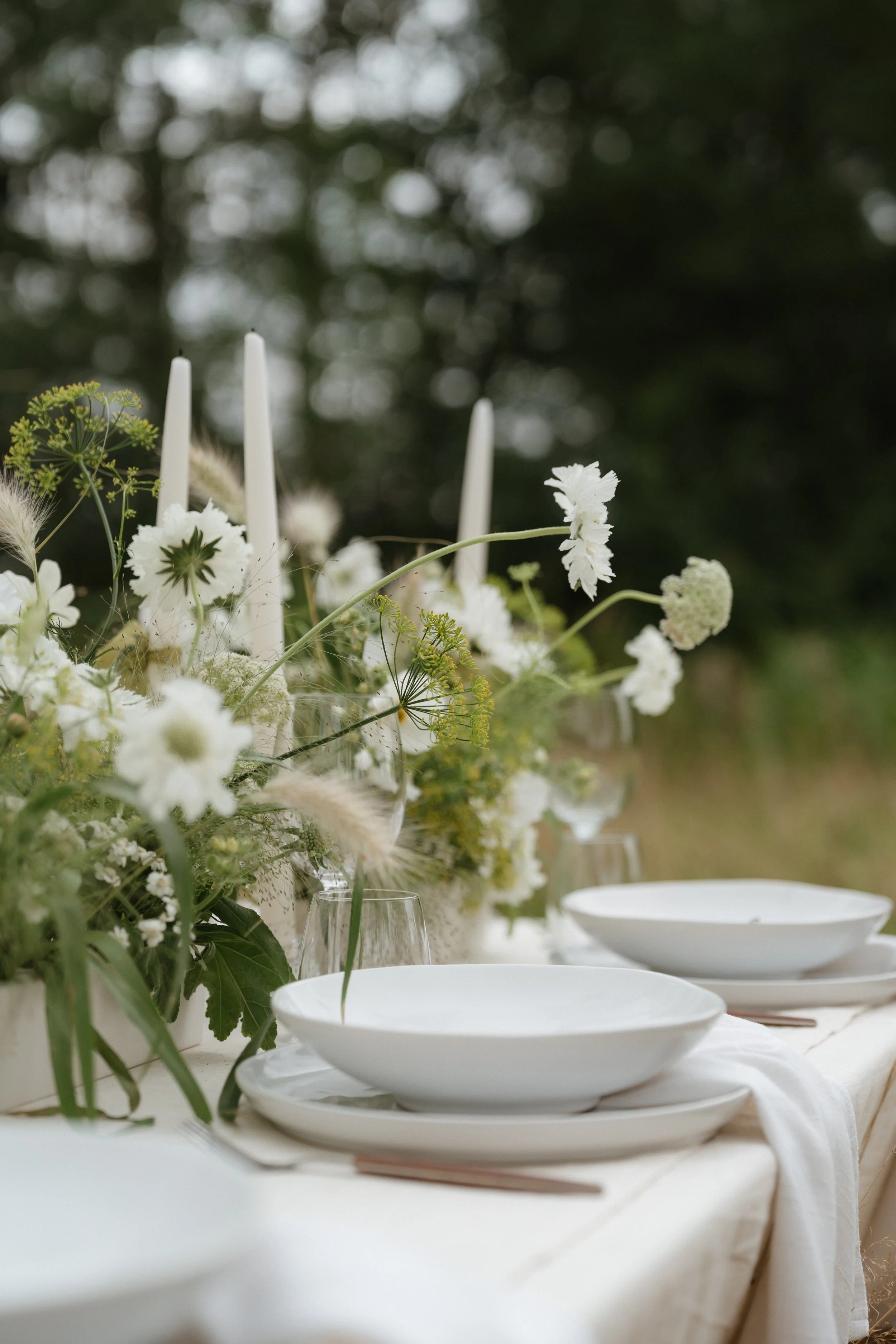 A table set outdoors with white plates, glasses, and a large floral centerpiece with white flowers and greenery. Candles in the background, dark greenery beyond.