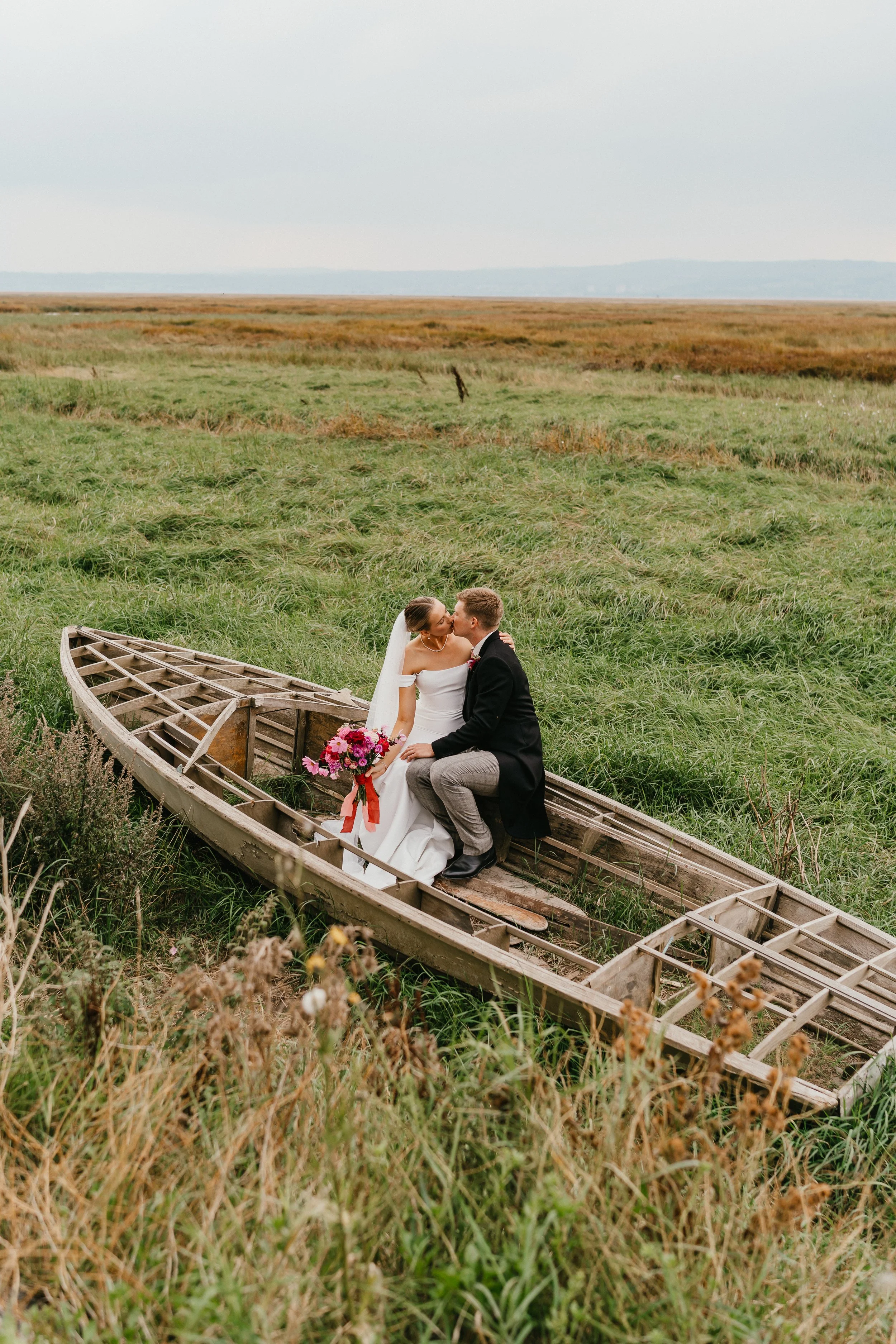 A bride and groom sitting in an old wooden boat on a grassy field on The Wirral, sharing a kiss. The bride is holding a colourful bouquet of flowers.