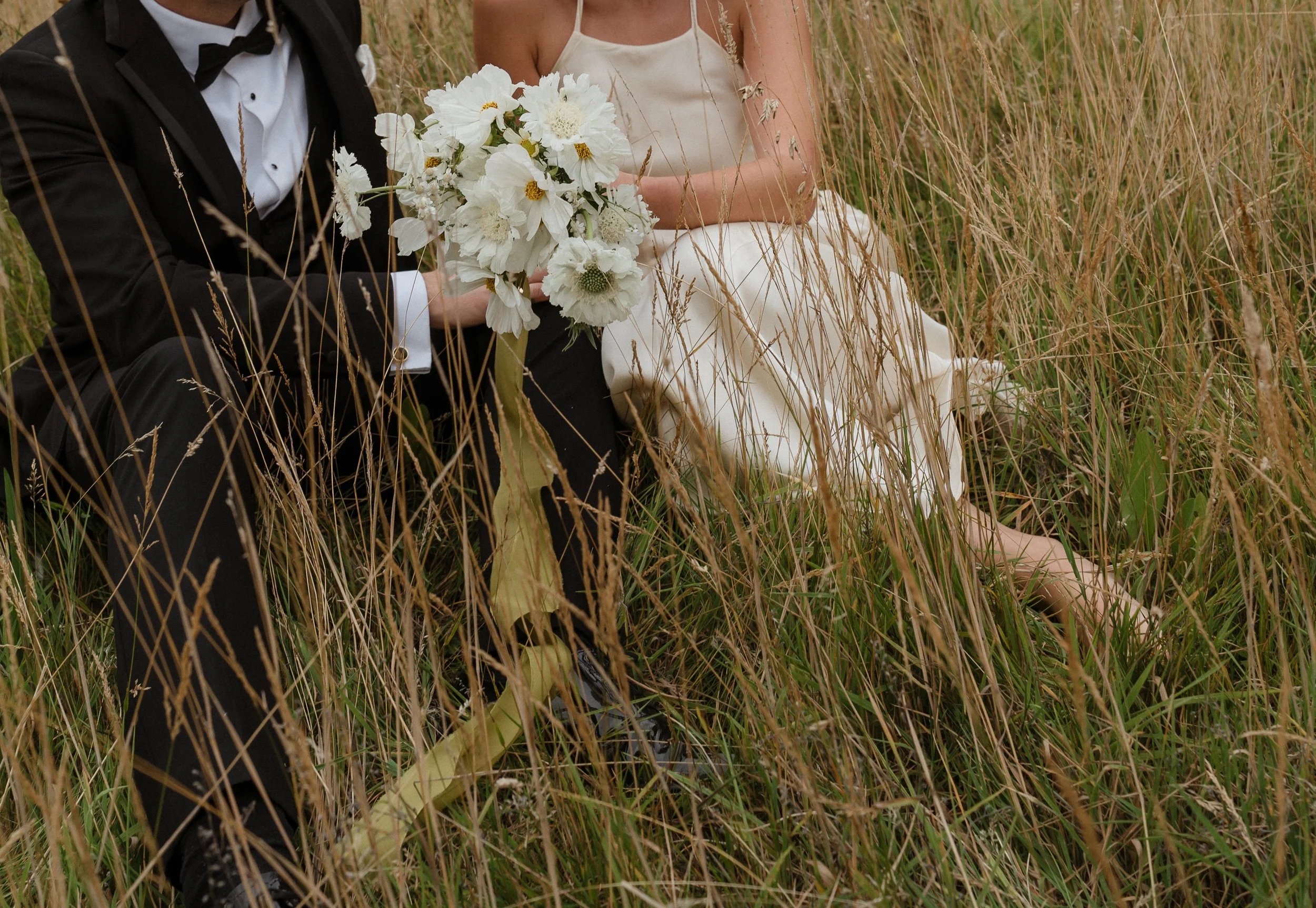 A couple sitting in tall grass, with the man wearing a tuxedo holding a bouquet of white flowers, and the woman in a white dress sitting beside him.