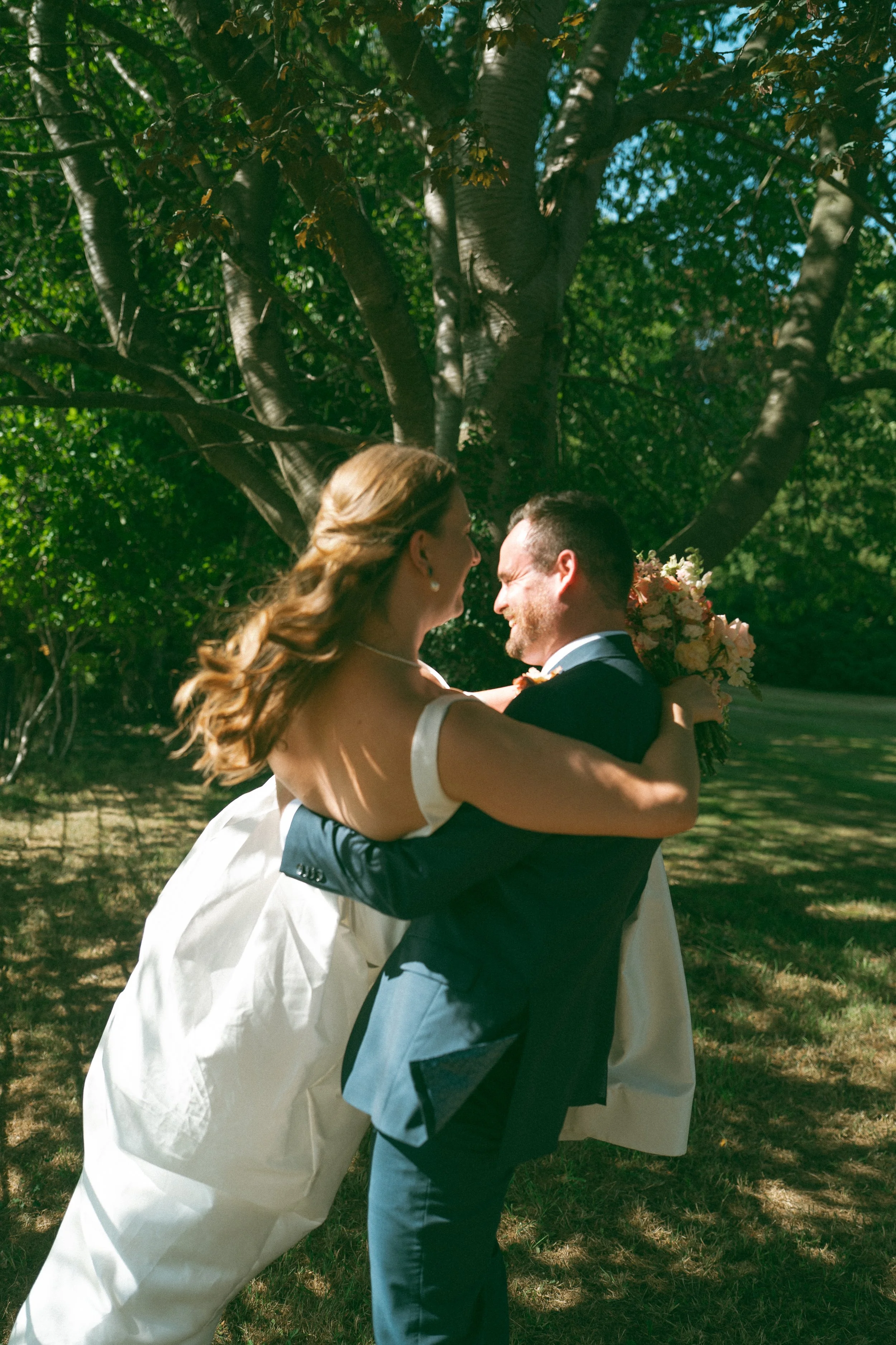 A bride and groom are dancing outdoors under a large tree at Goldsborough Hall, with the bride holding a bouquet of flowers and both smiling at each other.