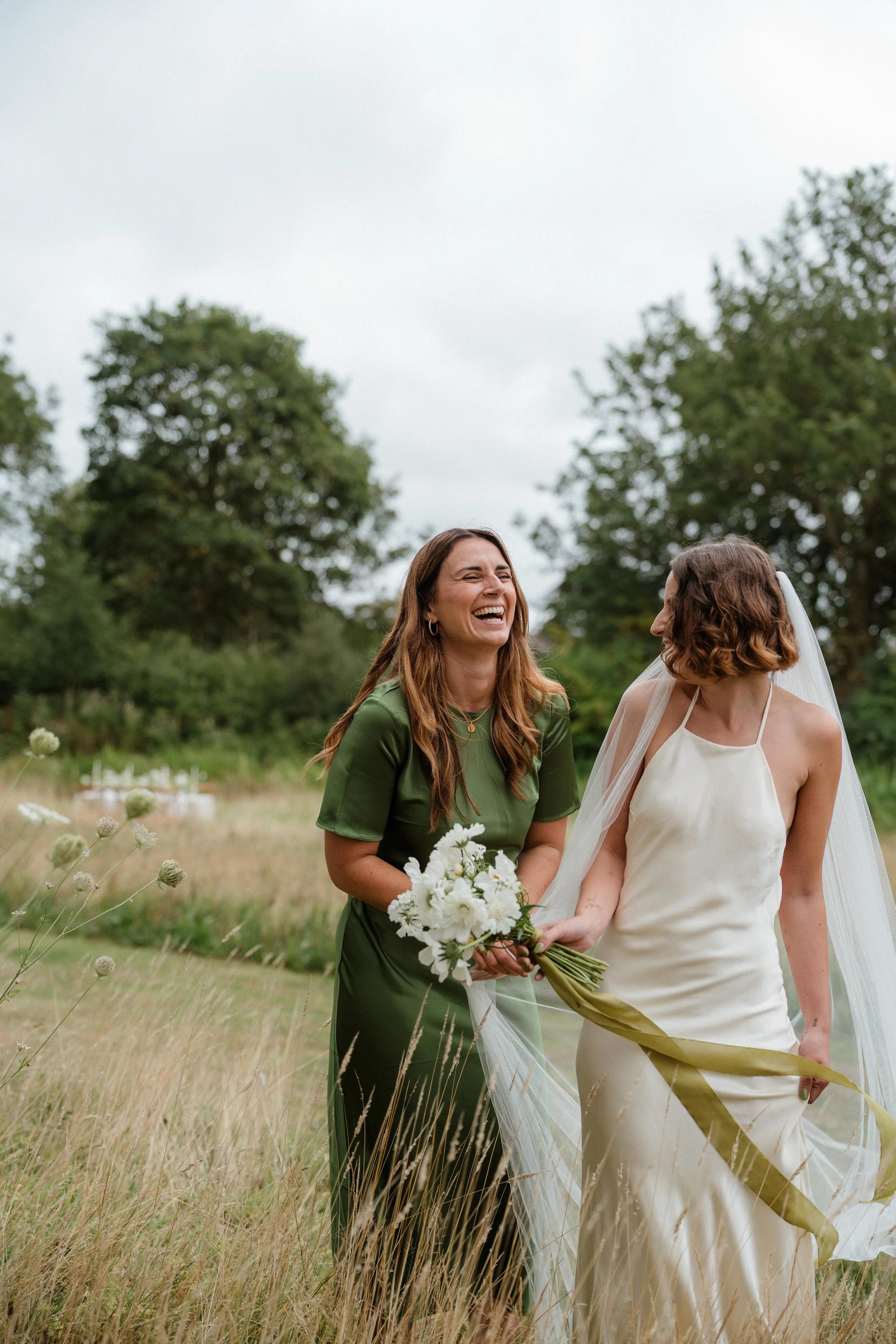 Two women, one in a white wedding dress and veil, and the other in a green dress, sharing a joyful moment outdoors in a grassy field with trees in the background, with one holding a bouquet of white flowers.
