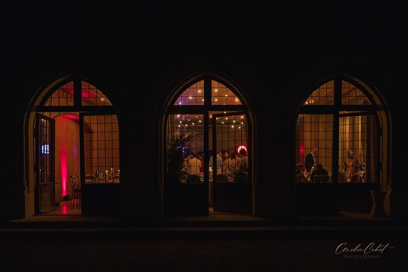 A photo from outside looking into the reception venue. Large windows frame the guests as fairy lights decorate the ceiling