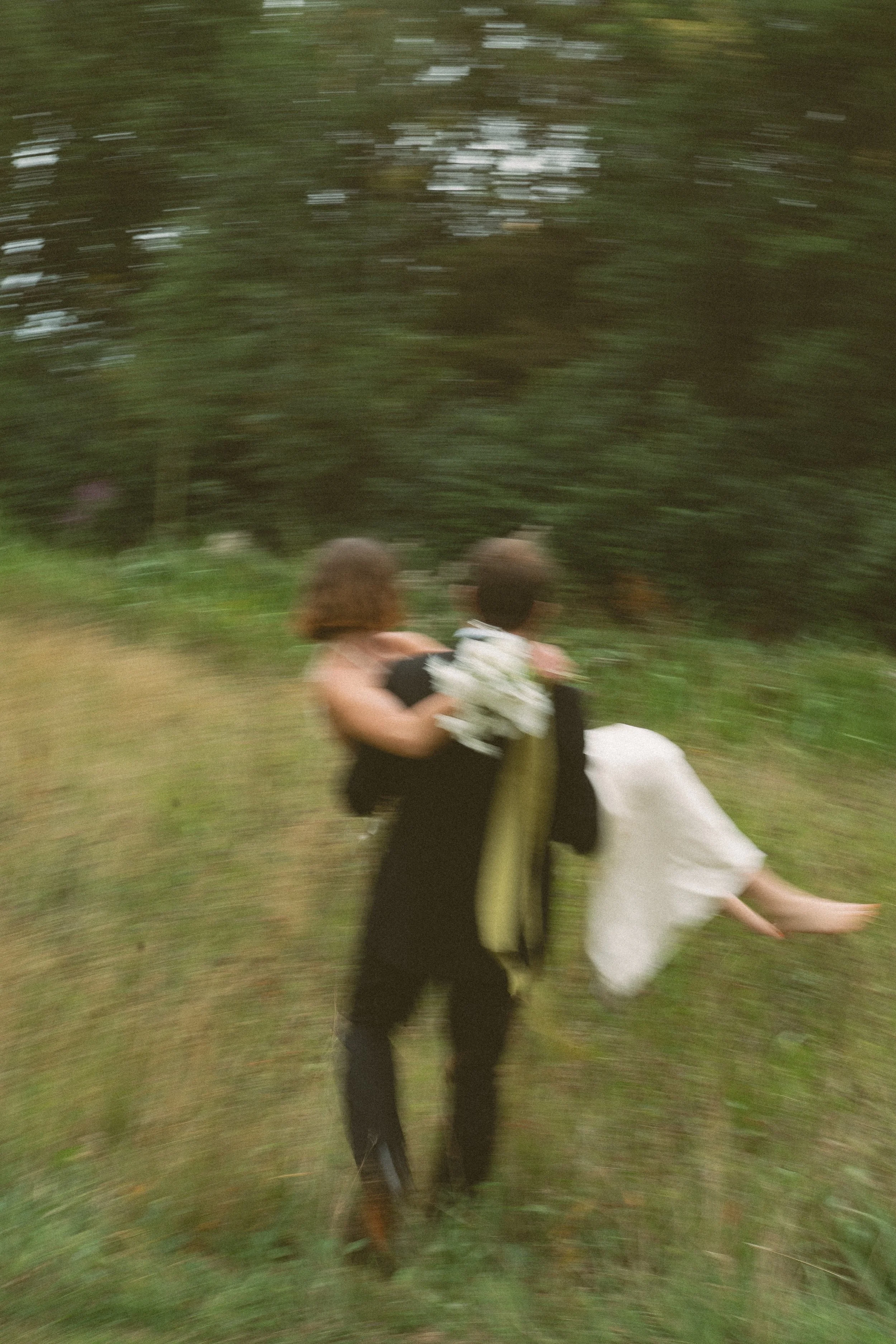 A man in a black suit carrying a woman in a white dress through a grassy area with trees in the background, motion blur.