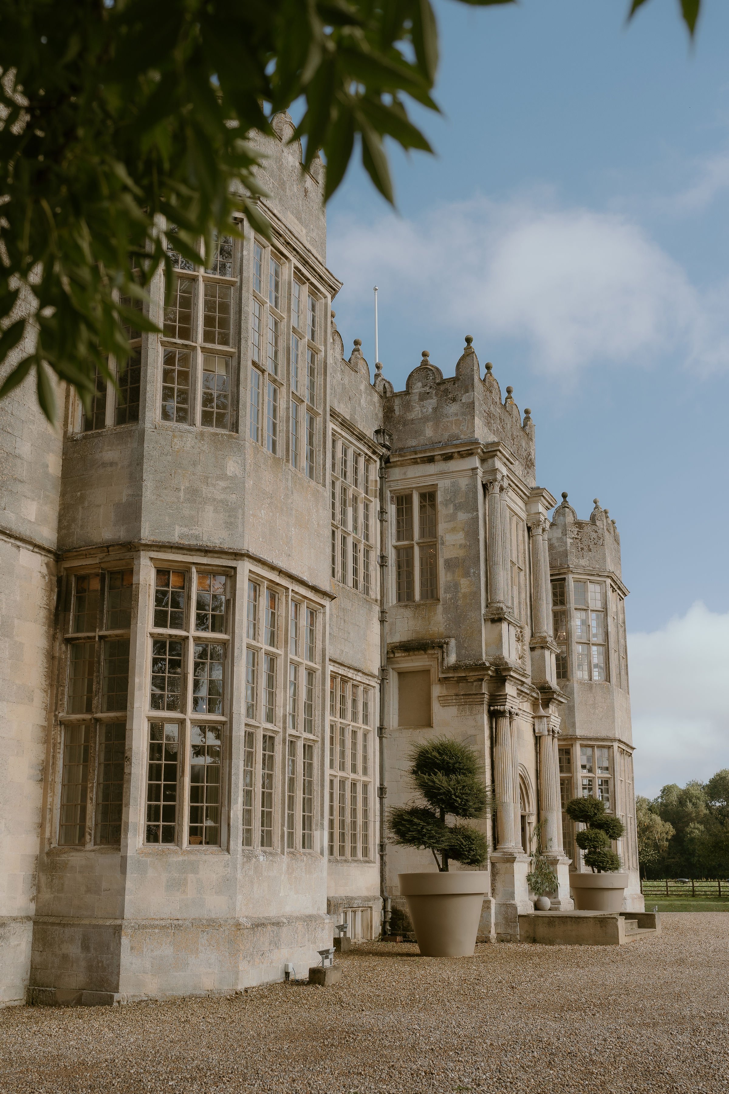 Front view of Howsham Hall - one of Yorkshire's top wedding venues - with large windows and potted plants at the entrance, under a partly cloudy sky.