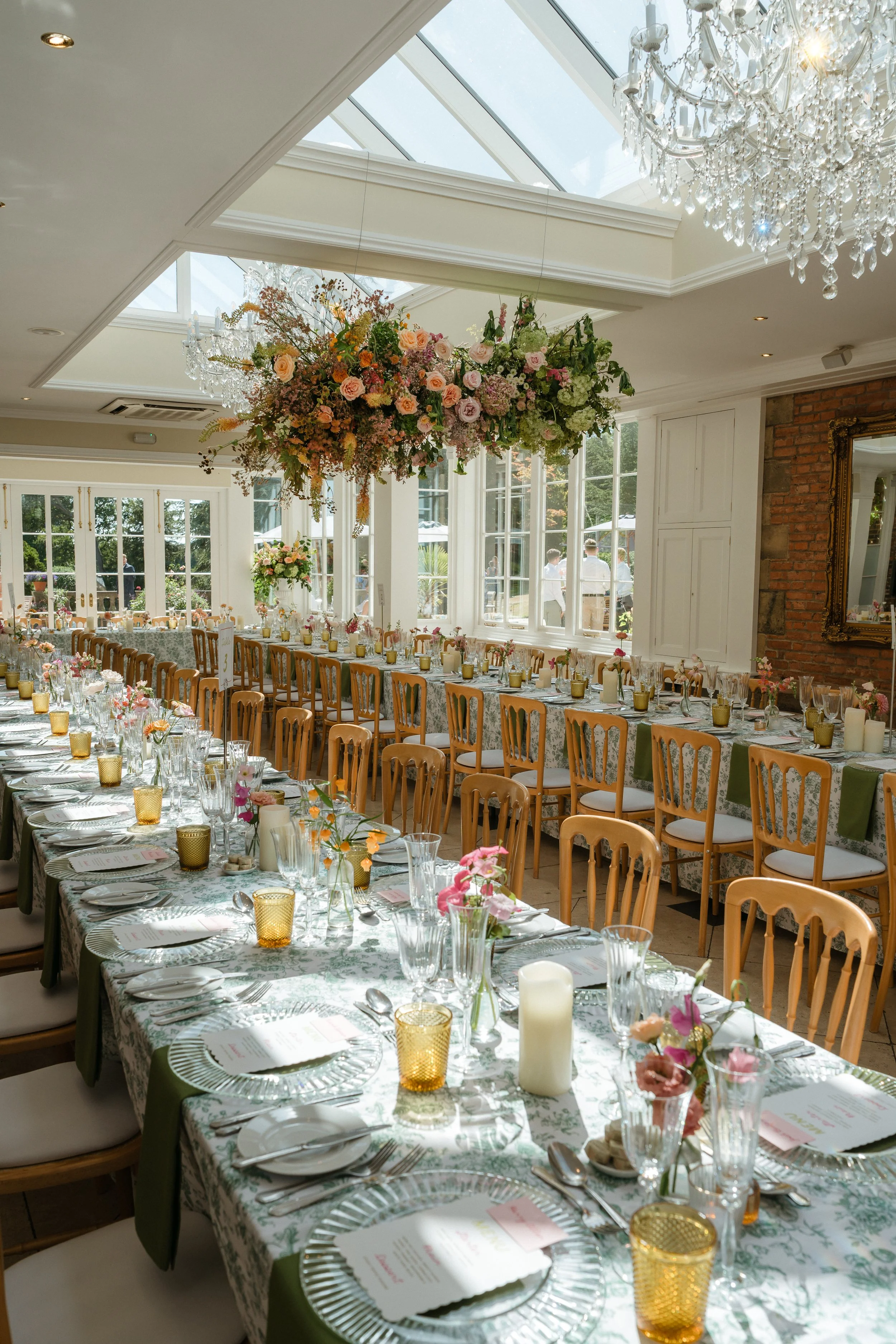 A long dining table set for a wedding at Goldsborough Hall, decorated with pink and white flowers, candles, glasses, and plates, inside a bright room with large windows and a glass ceiling, and a chandelier hanging from the ceiling.