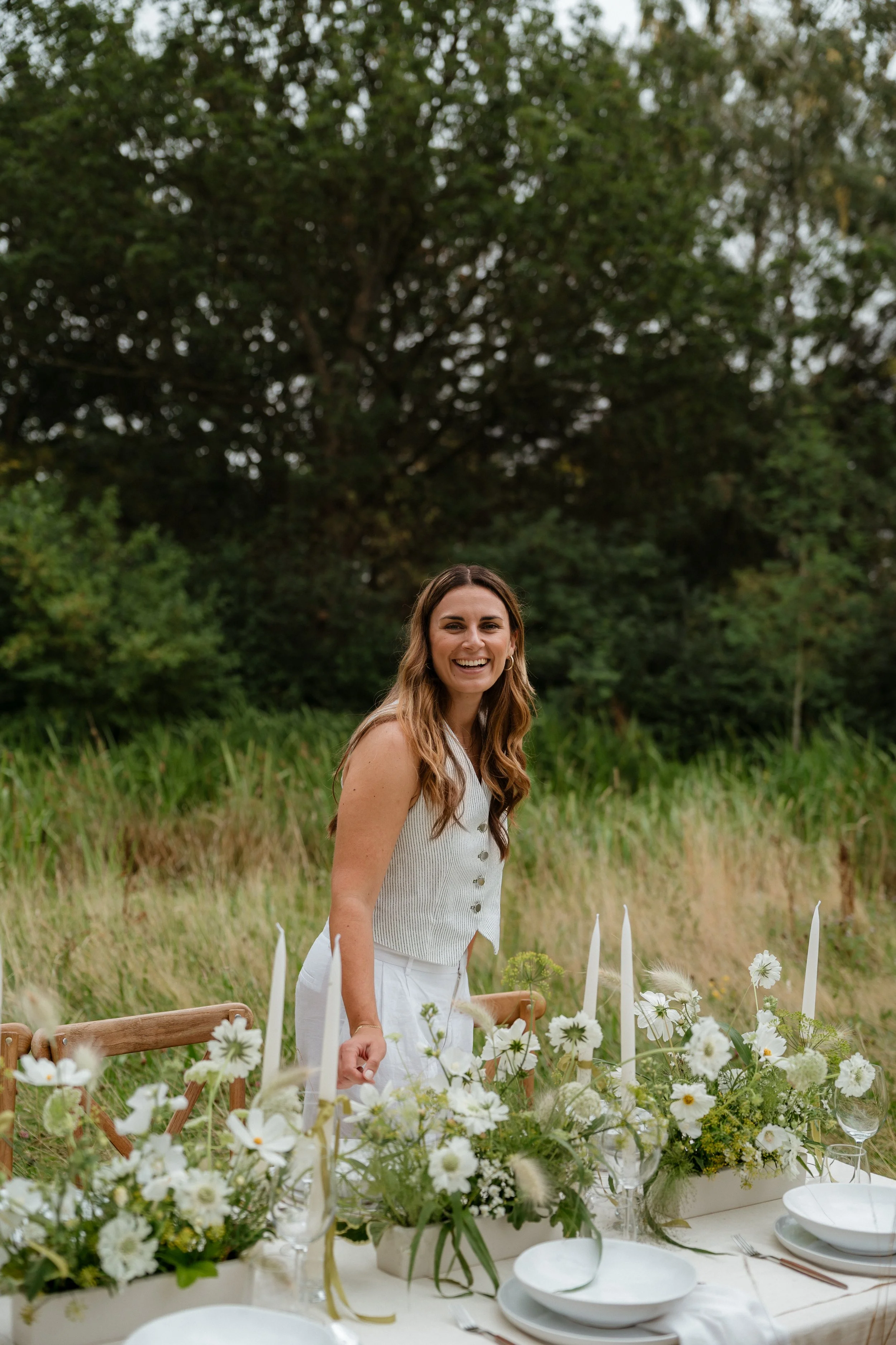 A woman smiling while standing next to a decorated outdoor dining table with white flowers and candles in a natural setting with greenery and trees in the background.