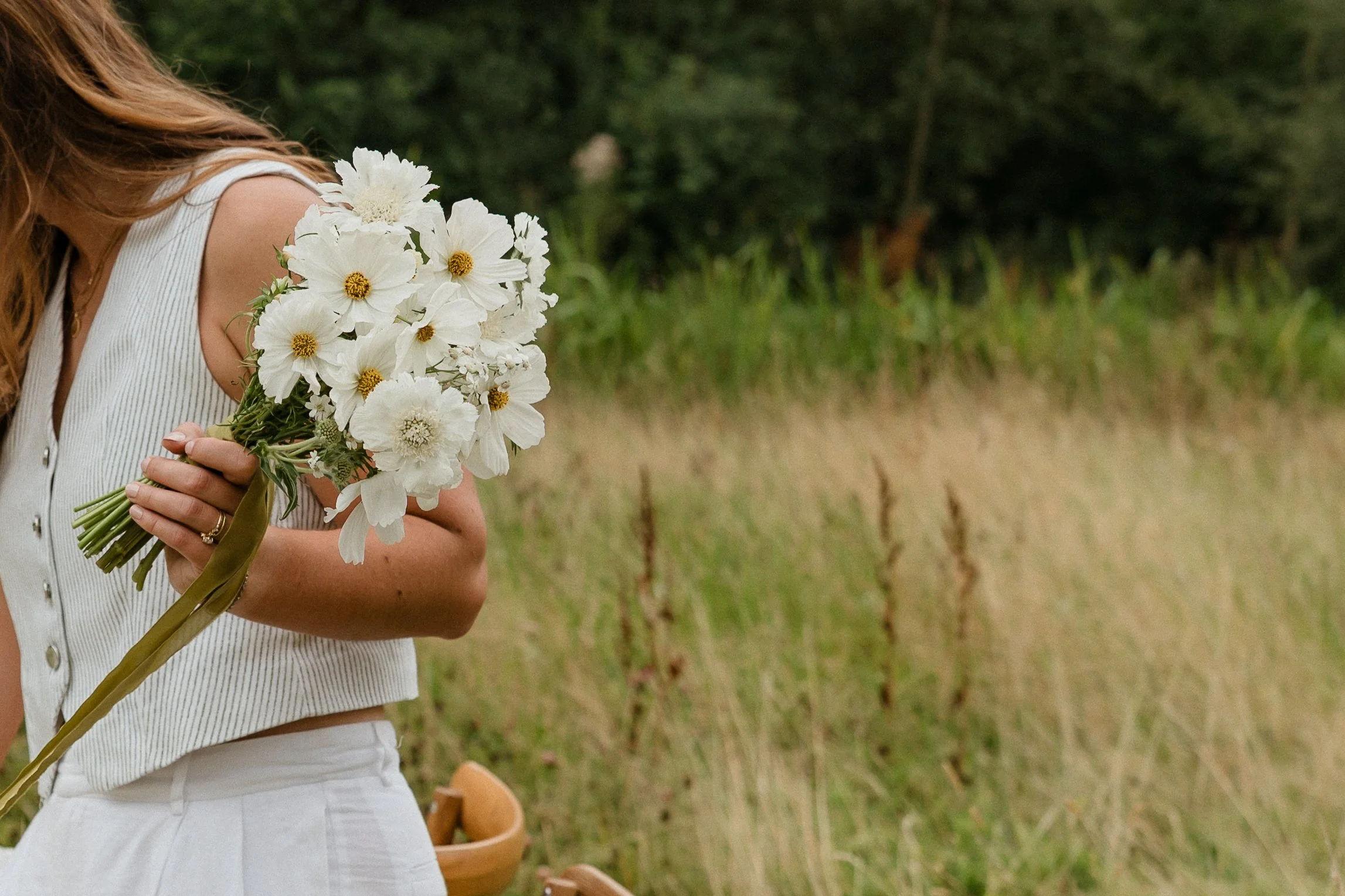 A woman holds a bouquet of white flowers with yellow centers in an outdoor setting with tall grasses and trees in the background.