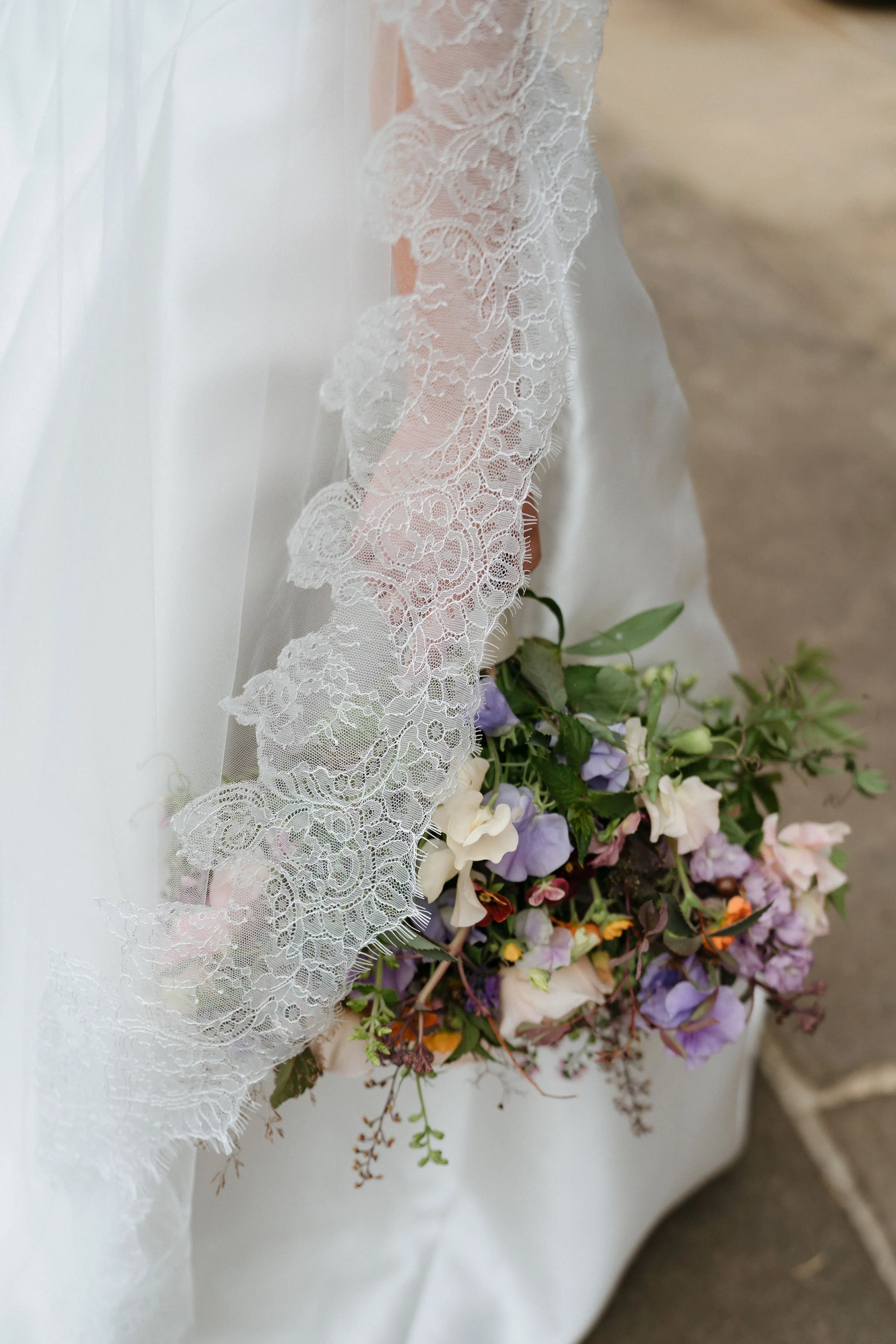 A white wedding dress with lace detail, holding a bouquet of pastel-colored flowers, on a white fabric surface.