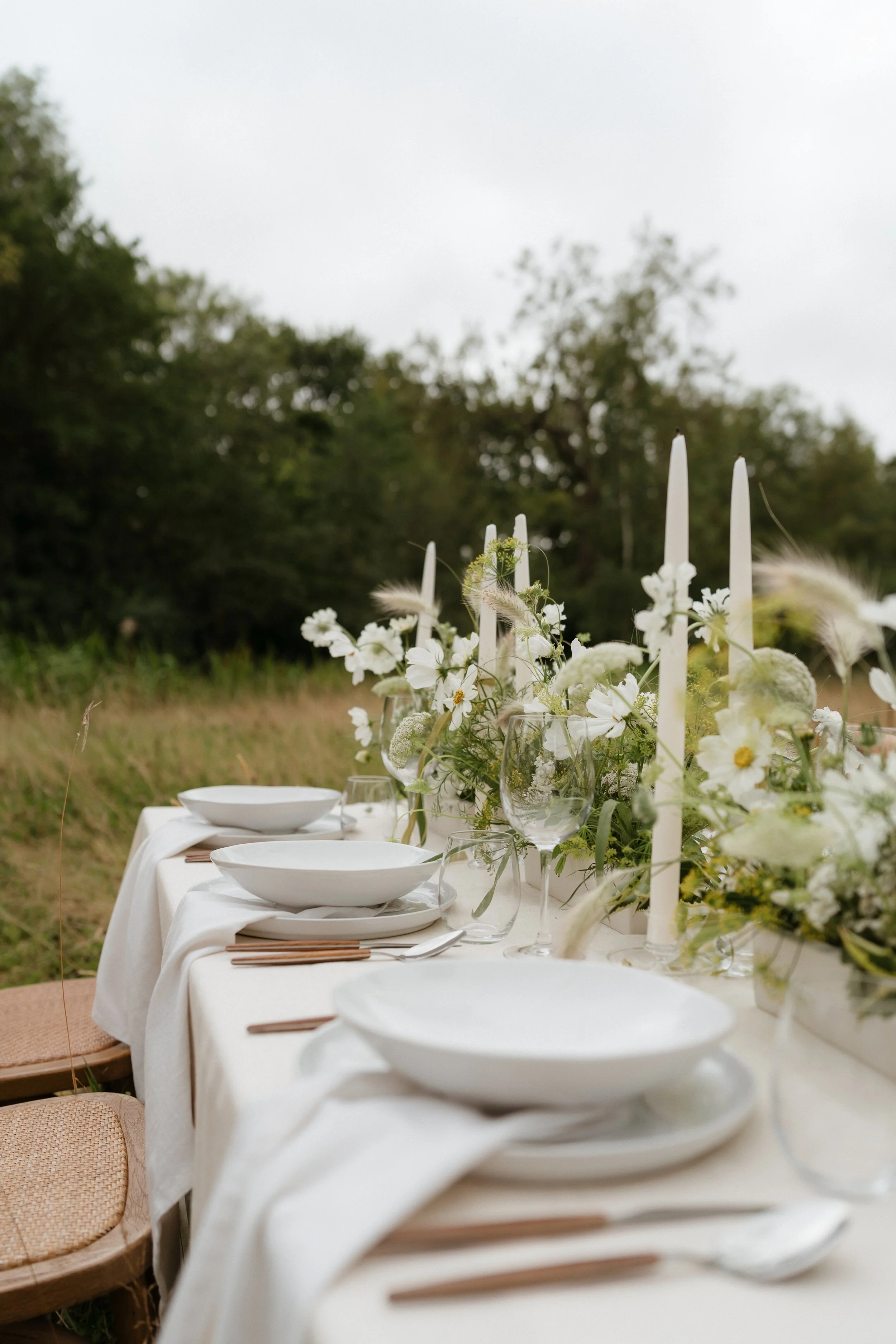 A long outdoor dining table set with white plates, glasses, and silverware, decorated with white flowers and tall white candles in a natural outdoor setting.