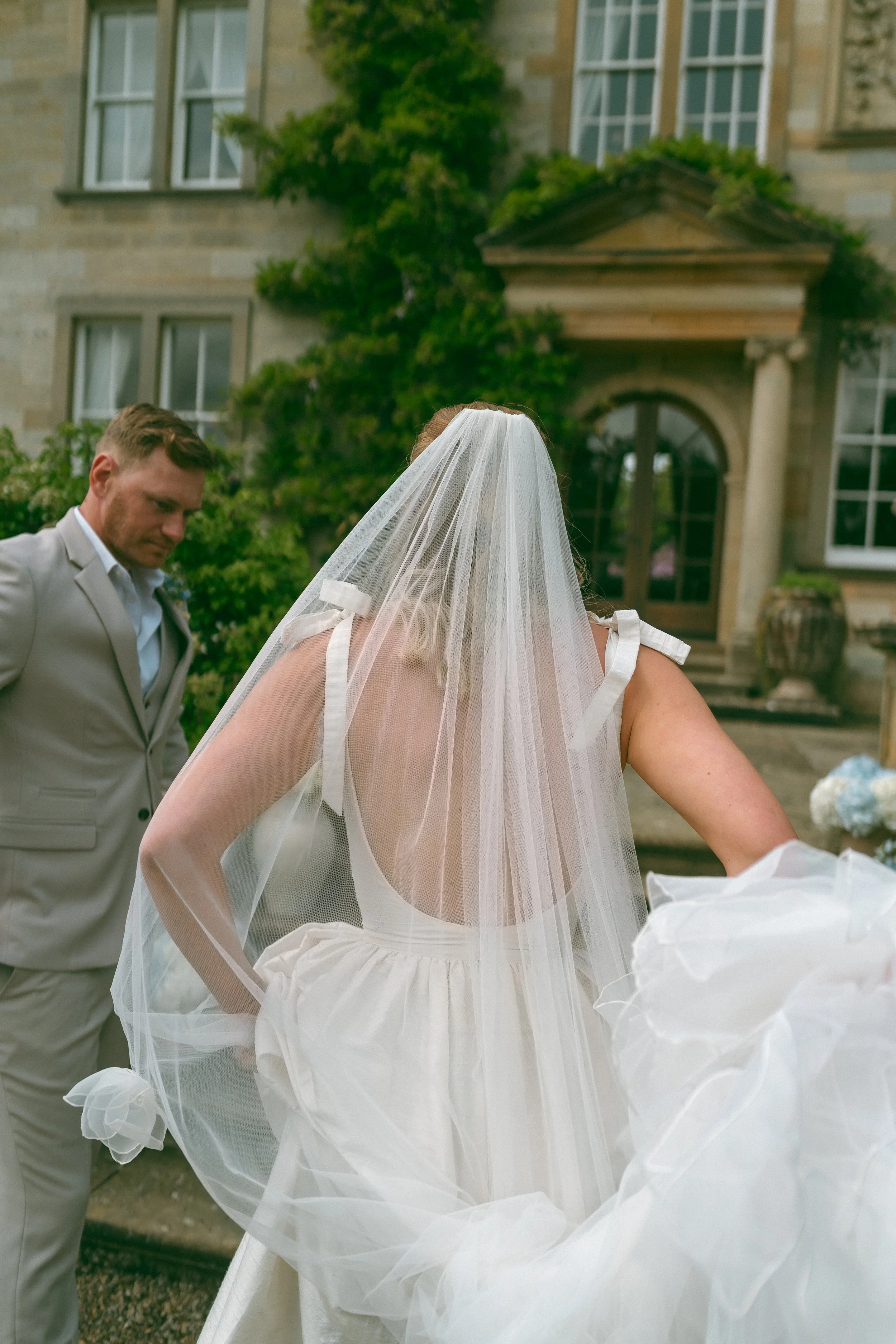 Bride wearing a white dress and veil with a man in a gray suit standing nearby outside a large house.