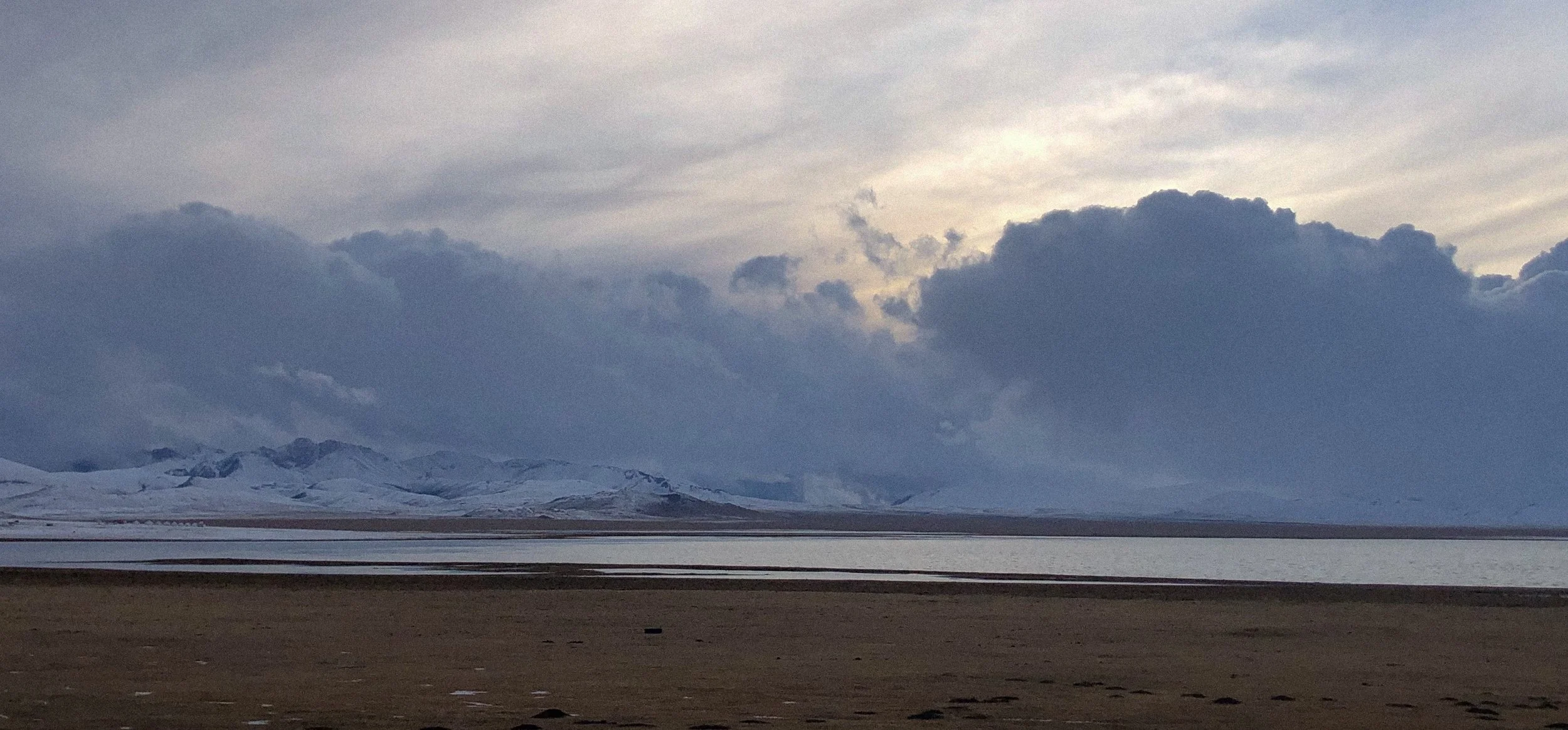 Cloudy sky over snow-capped mountains near a body of water and a sandy shoreline.