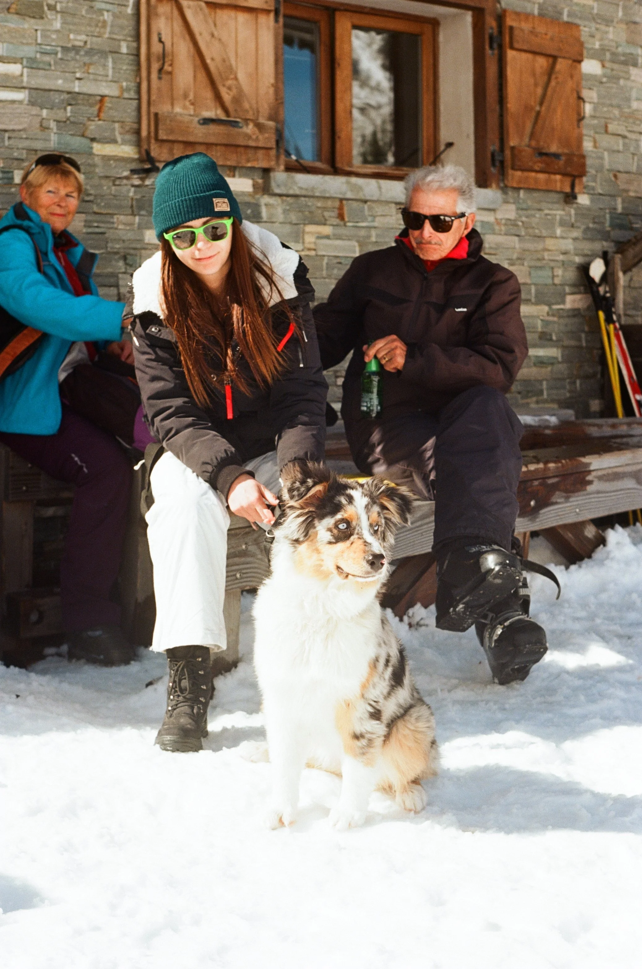 Three people sitting outside a stone cabin on snow, with a dog in the foreground. The woman in the middle is adjusting the dog's collar, the elderly man to her right is holding a drink, and a woman in a blue jacket is sitting behind them.