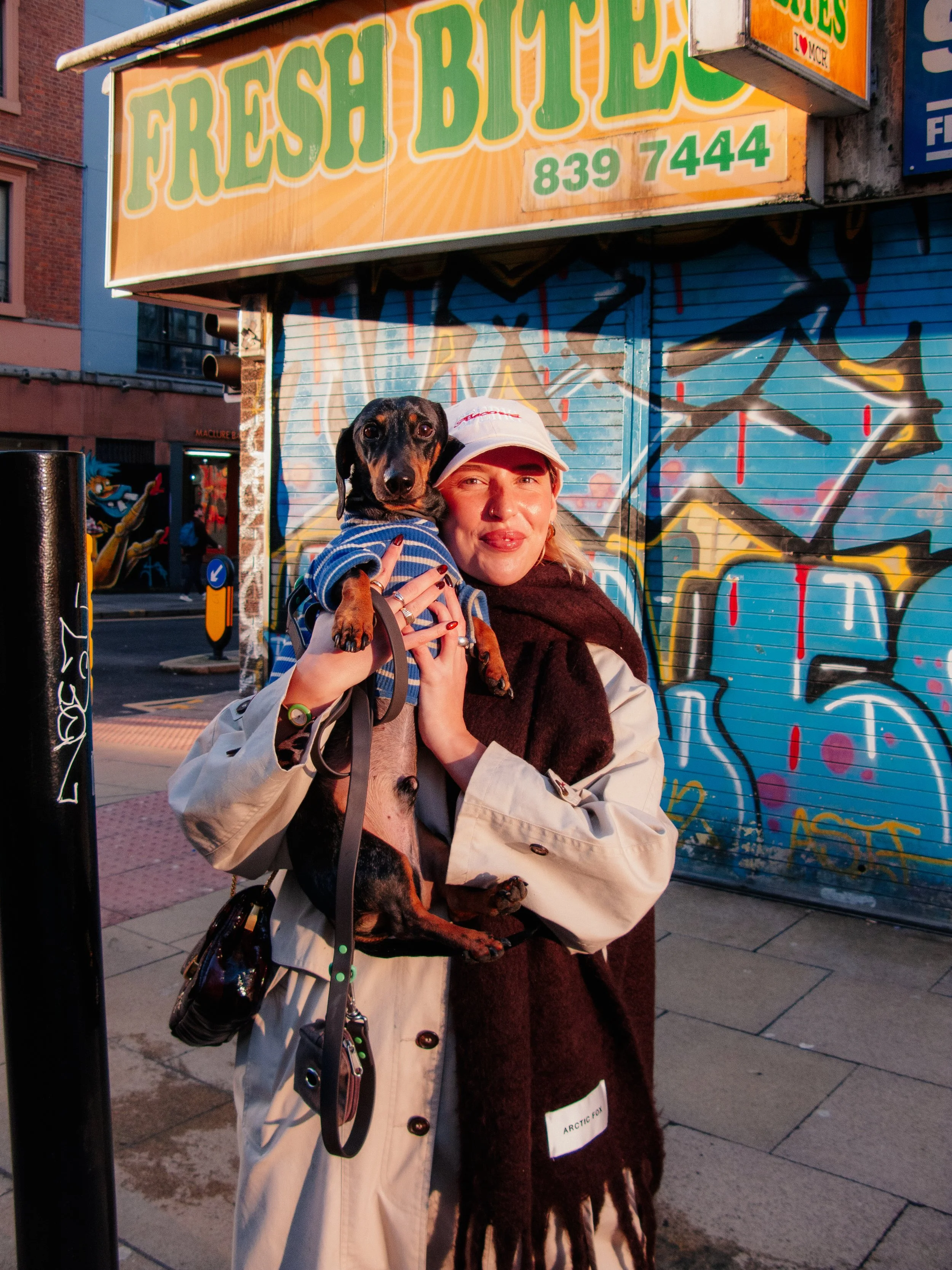 A woman holding a small black and tan dachshund dog in front of a colorful graffiti wall and a shutter with a bright yellow 'FRESH BITES' sign, on a city sidewalk.