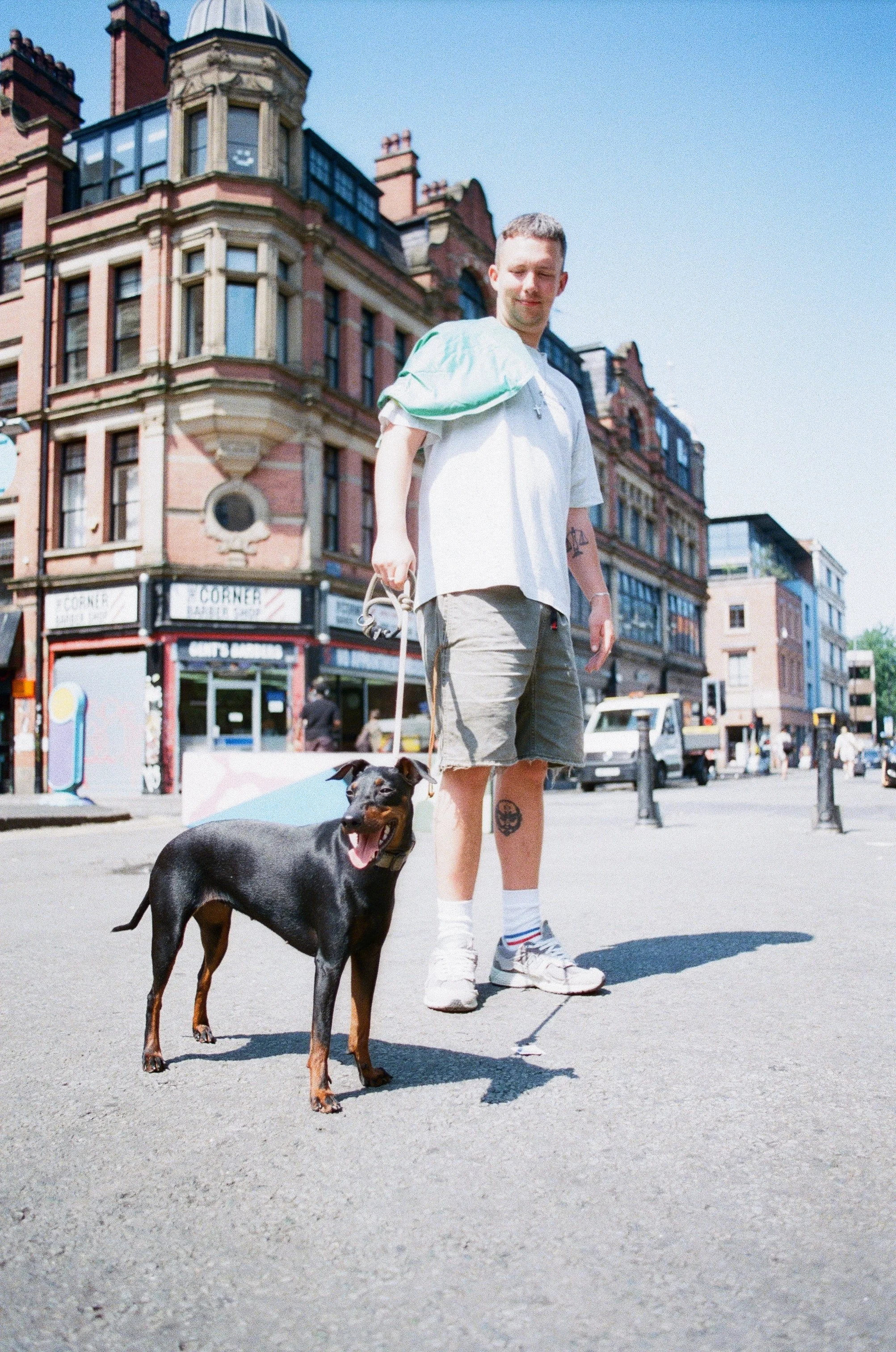 A man walking a black and tan dog on a leash in an urban street scene with historic buildings in the background on a sunny day.