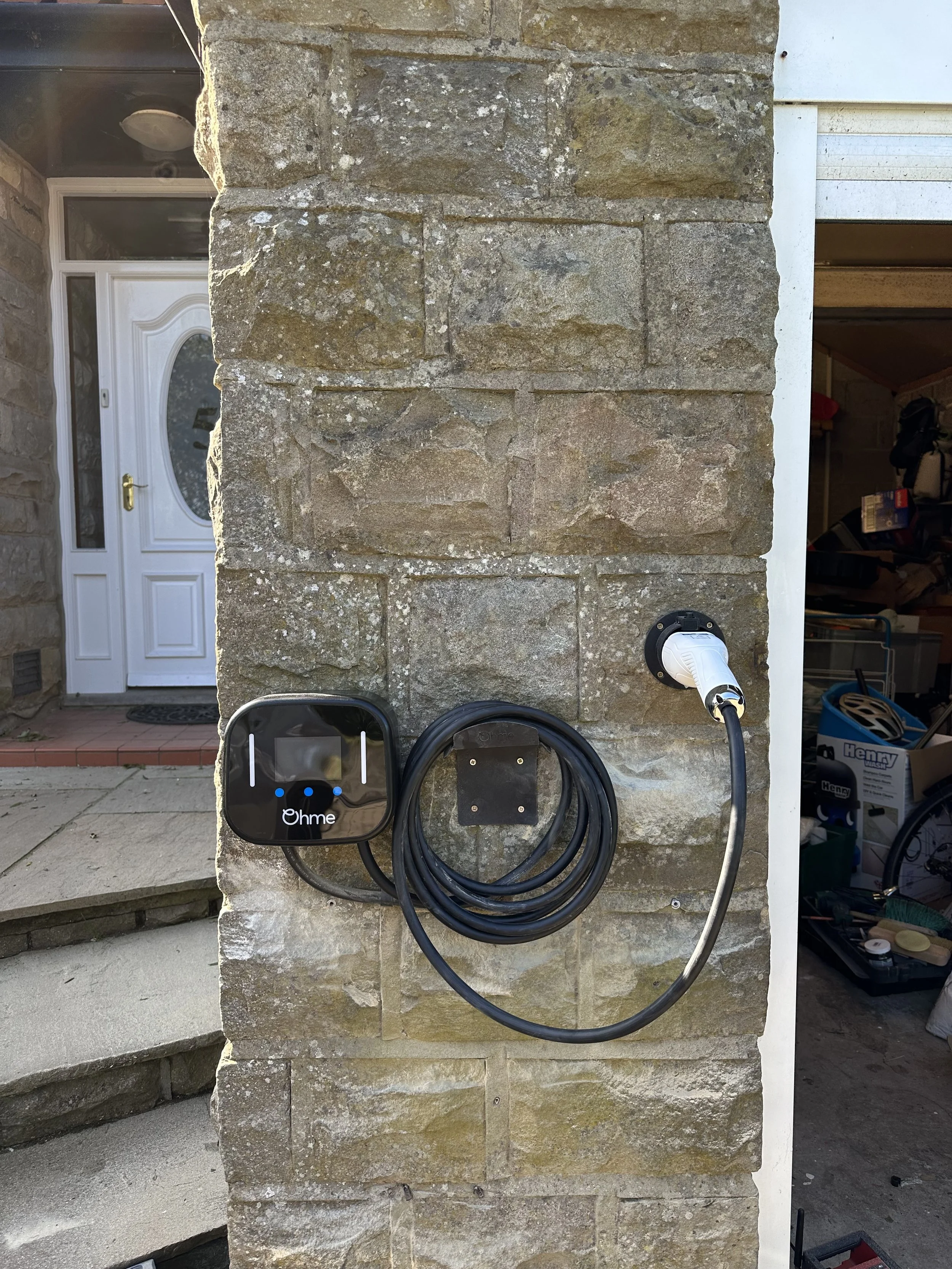 Electric vehicle charging station on a stone wall outside a house, with a black charging unit, a coiled cable, and a white charging plug connected to an outlet.