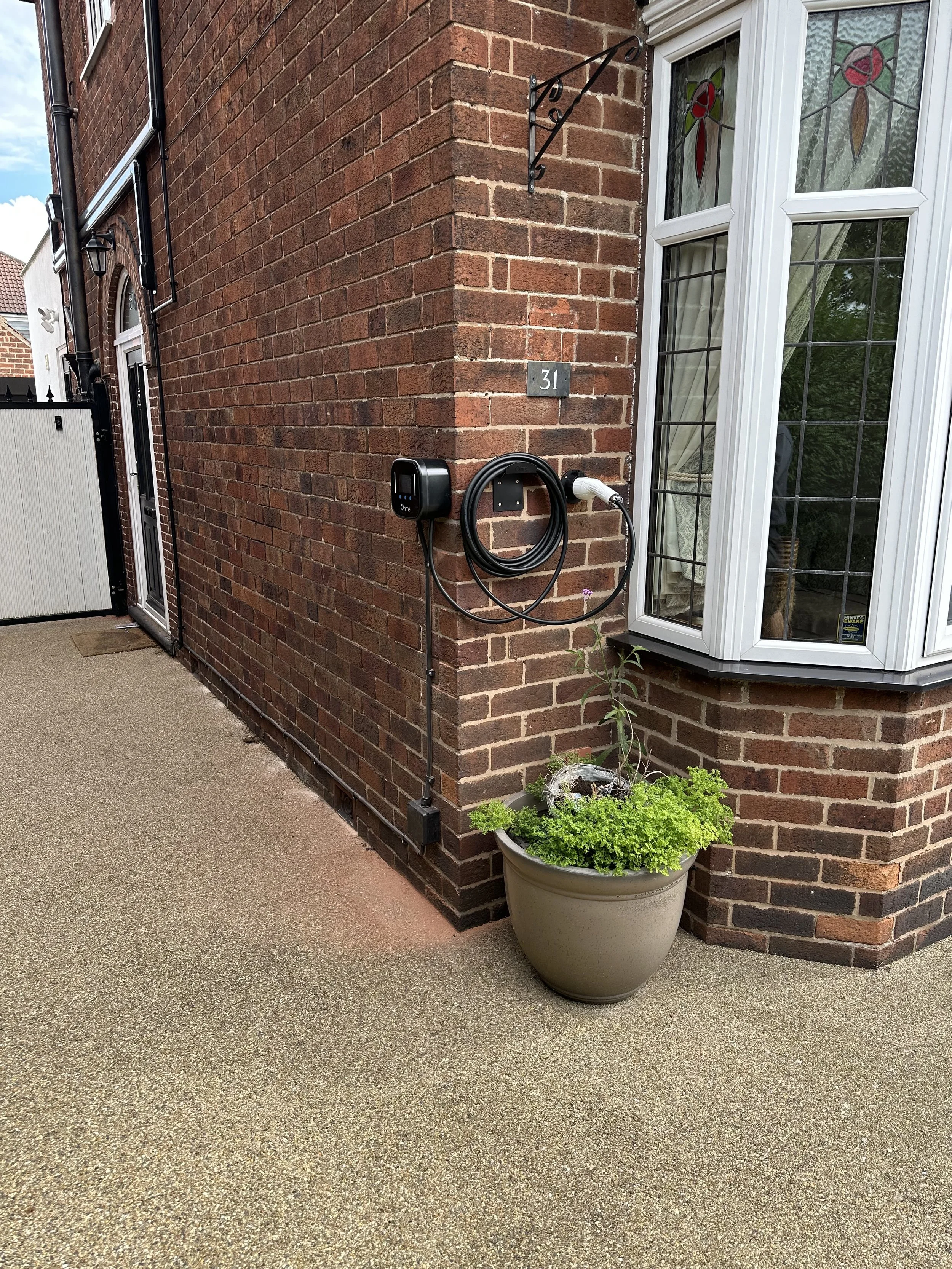 Exterior corner of a brick house with an electric vehicle charging station mounted on the wall, a potted plant below it, and a bay window with stained glass panels.