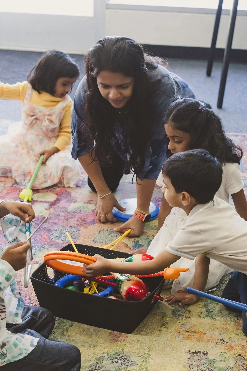 A mother and children sitting on a rug choose rhythm instruments from a basket.
