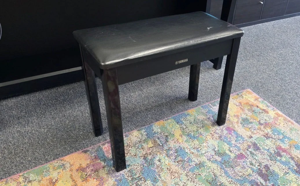 A standard, non-adjustable black piano bench with some cosmetic wear and padded seating.