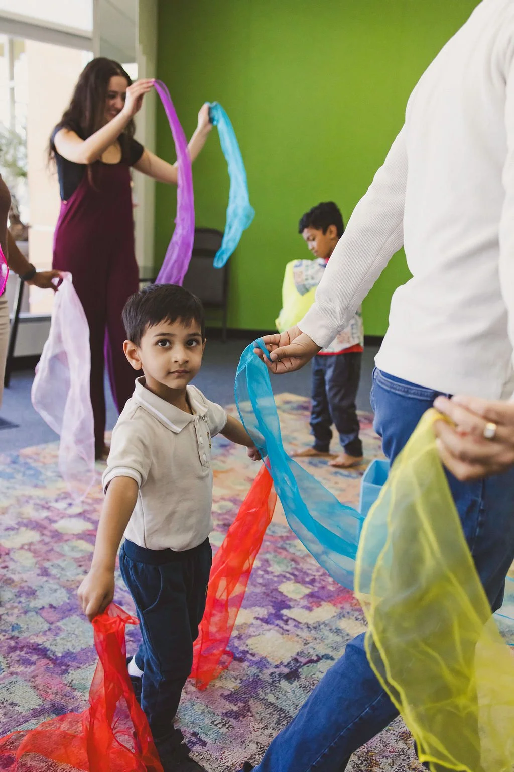 A music class of children and parents move around the room while holding long, brightly-colored scarves.