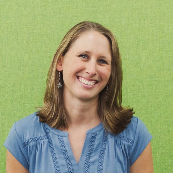 A woman with light brown hair, wearing a blue top, stands in front of a green background 