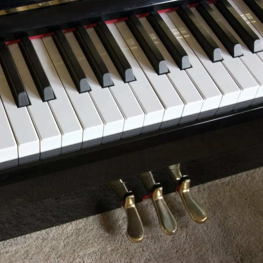 A zoomed-in image of a high-gloss, black piano. The keys and three gold-toned pedals are both visible.