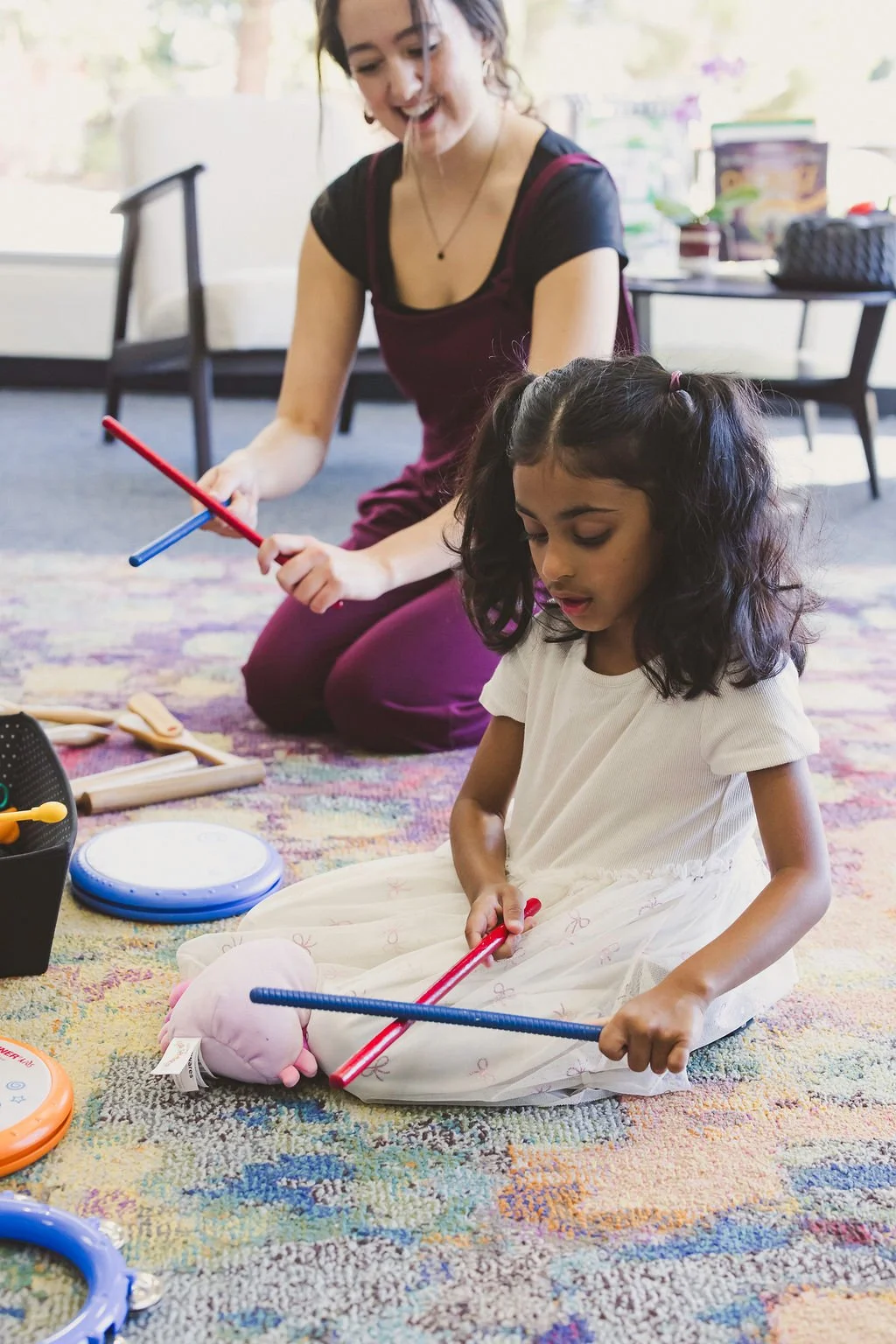 A young child sits on a rug, tapping two rhythm sticks together. The music teacher sits in the background with her own rhythm sticks.
