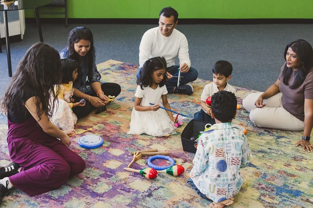 A class of children and parents sit on a large rug, playing rhythm instruments