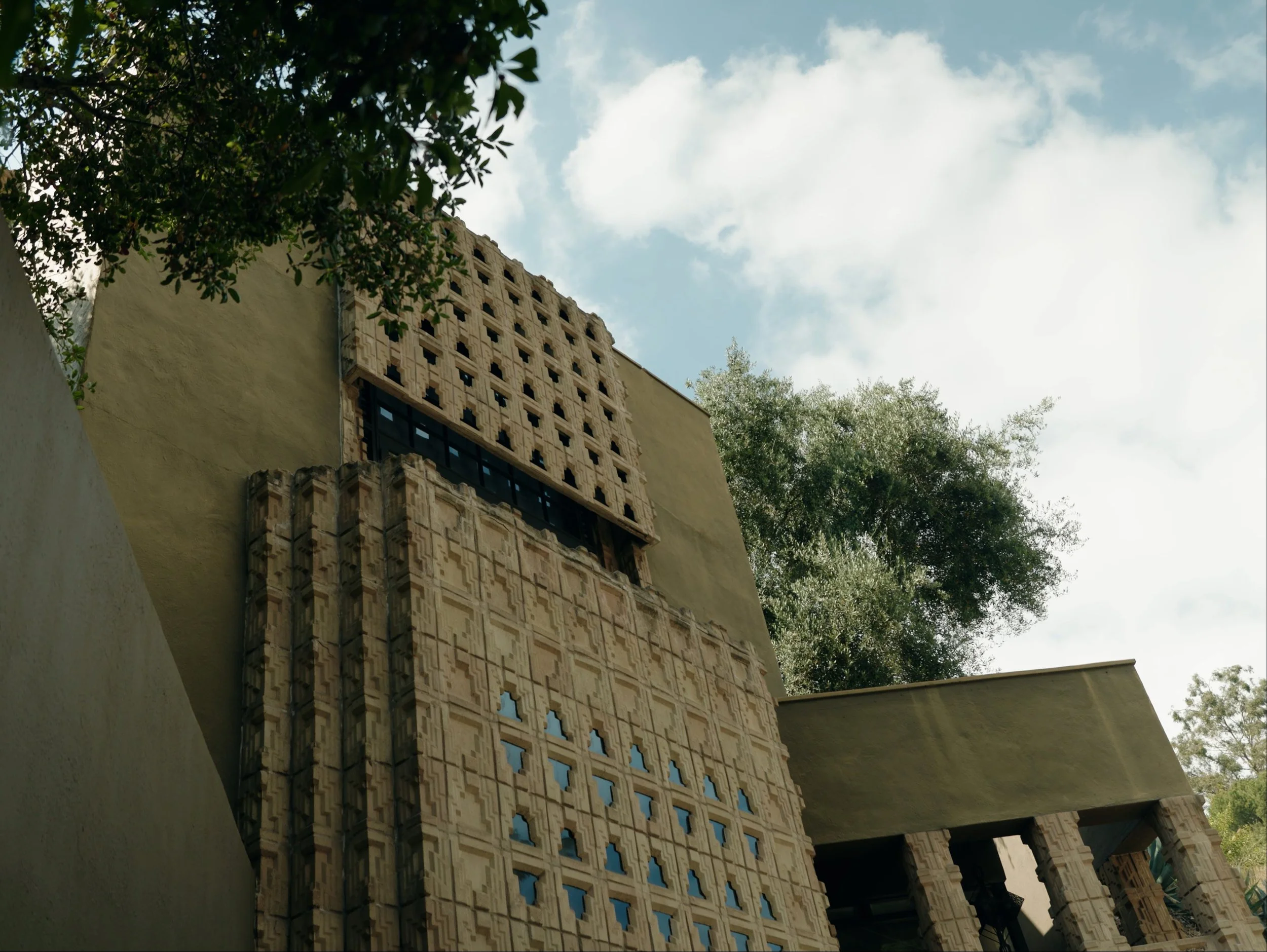 An architectural detail of a building with intricate patterned brickwork and small blue window openings, surrounded by leafy green trees, against a partly cloudy sky.