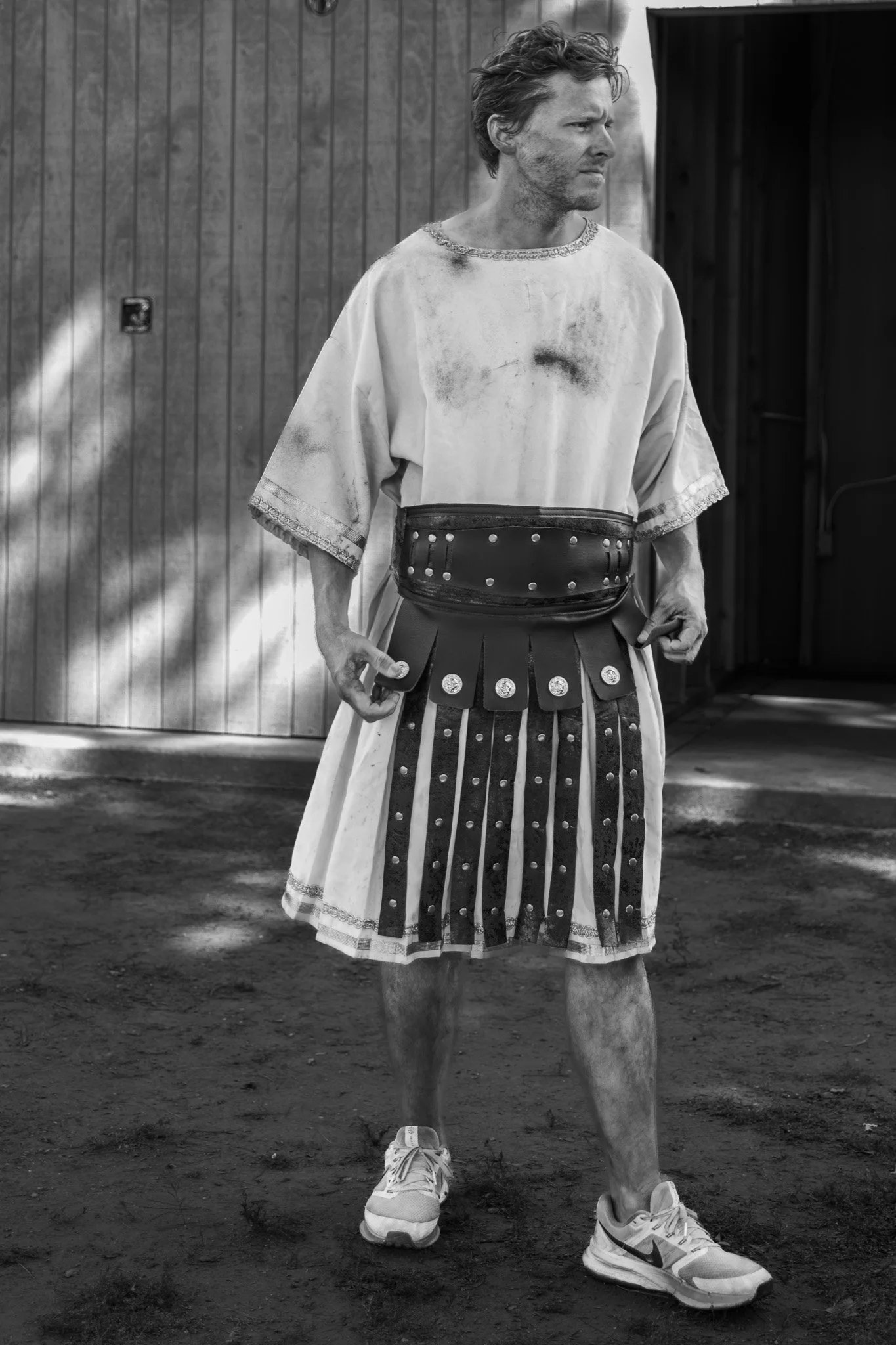 A man standing outdoors on a dirt surface, wearing a medieval-style costume with a tunic, a wide belt with metal accents, a skirt with patterns, and modern sneakers, while looking to the side with a serious expression.