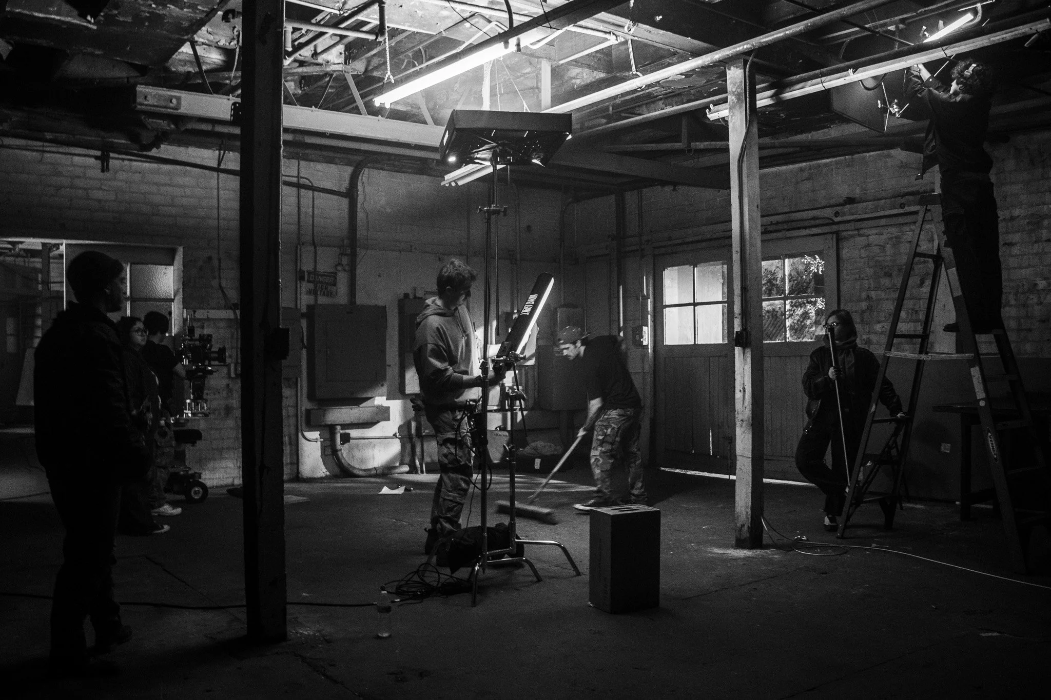 Black and white photo of a film set inside a garage or warehouse. Crew members operate equipment, including a boom microphone and lighting. A person on a ladder adjusts a light. Large windows and visible pipes are in the background.
