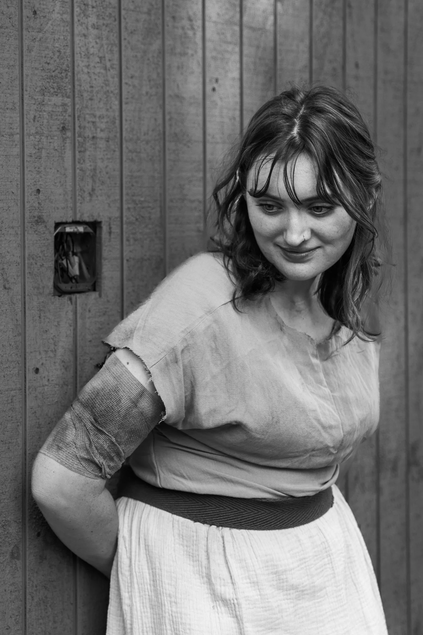 A young woman with styled wavy hair, wearing a light-colored top and a belt, stands against a wooden fence background, looking down slightly with a subtle smile.