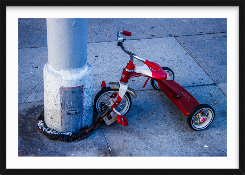 Child's red and white tricycle chained to a concrete lamp post on a sidewalk.