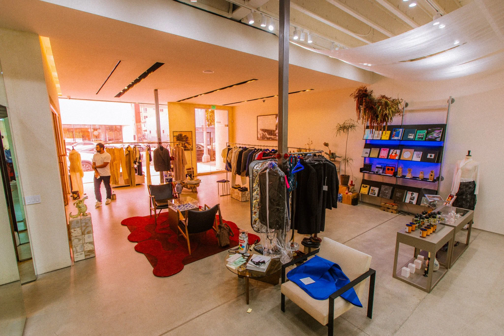Inside a retail store displaying clothing on racks and shelves, with a man browsing, and a window at the front letting in natural light.